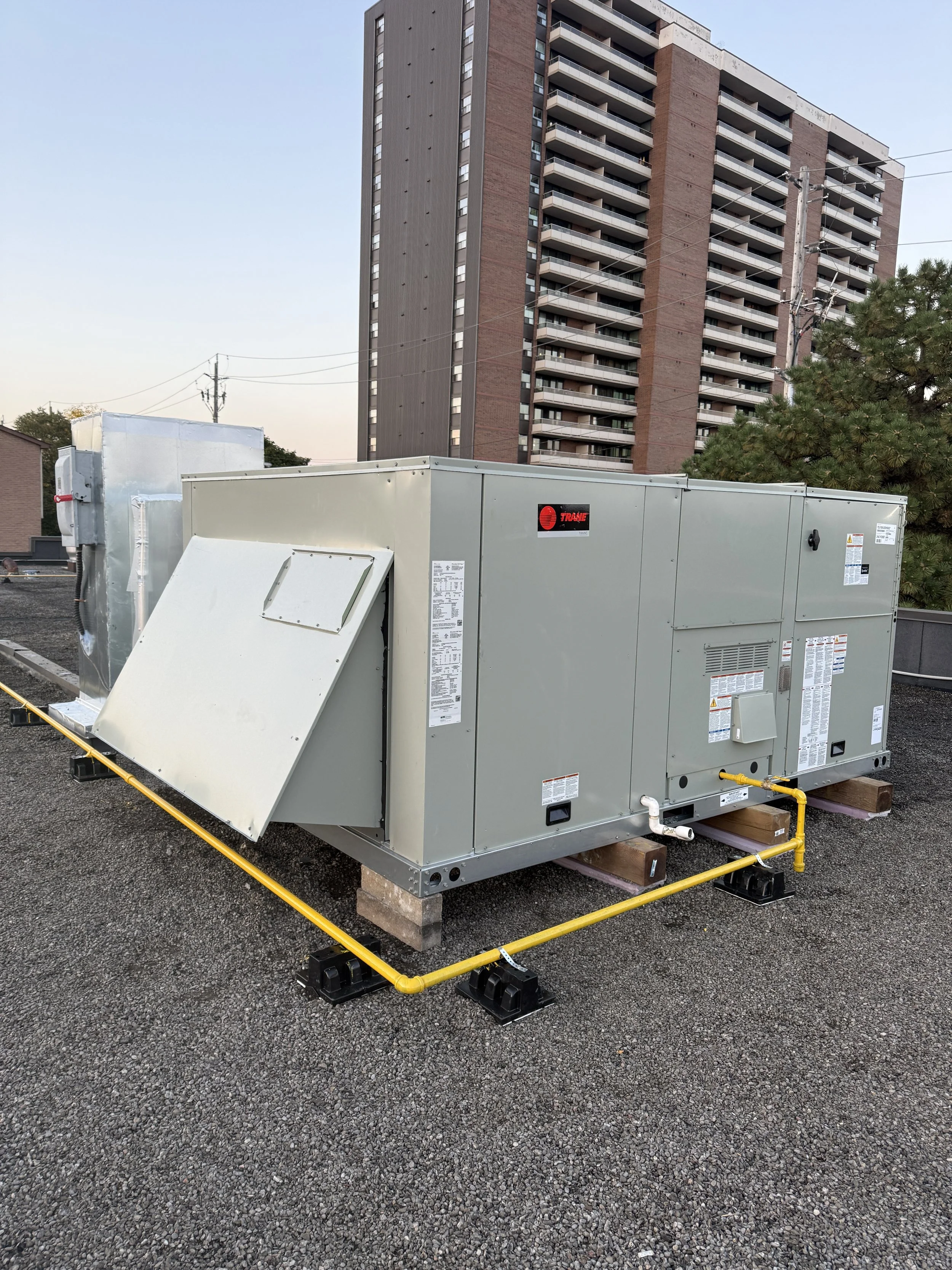 Commercial rooftop HVAC unit with yellow gas line, in front of a high-rise apartment building, on a gravel surface.
