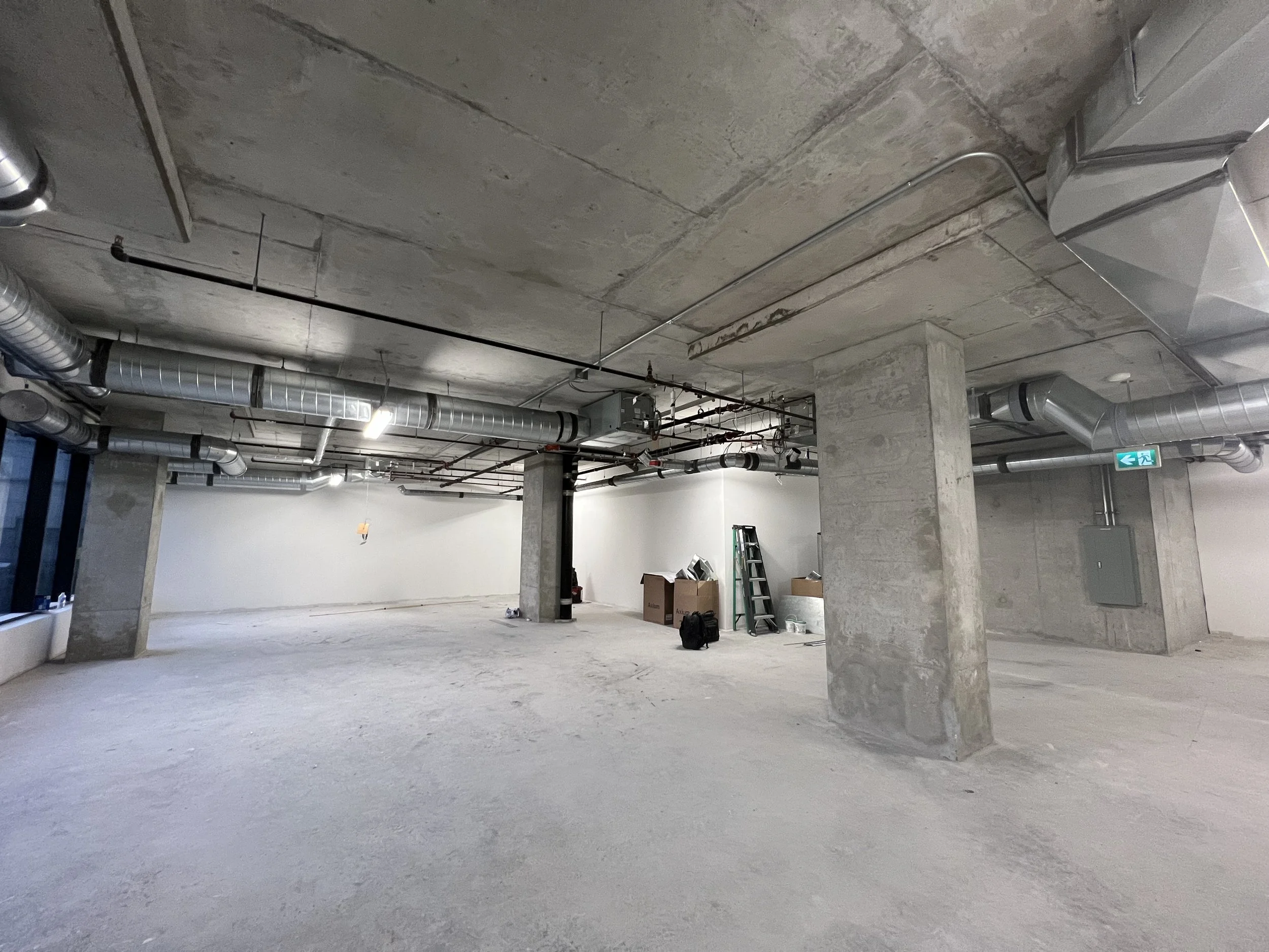 An empty unfinished interior space with exposed concrete ceiling and floor, metal air ducts, a ladder, boxes, and an emergency exit sign.