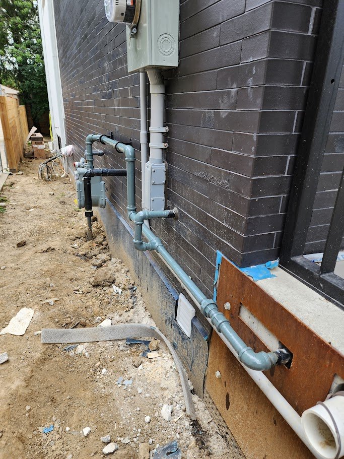 The image shows building plumbing and electrical work with pipes, a meter, and electrical boxes installed along the exterior brick wall of a house, near a window and an unfinished dirt ground.