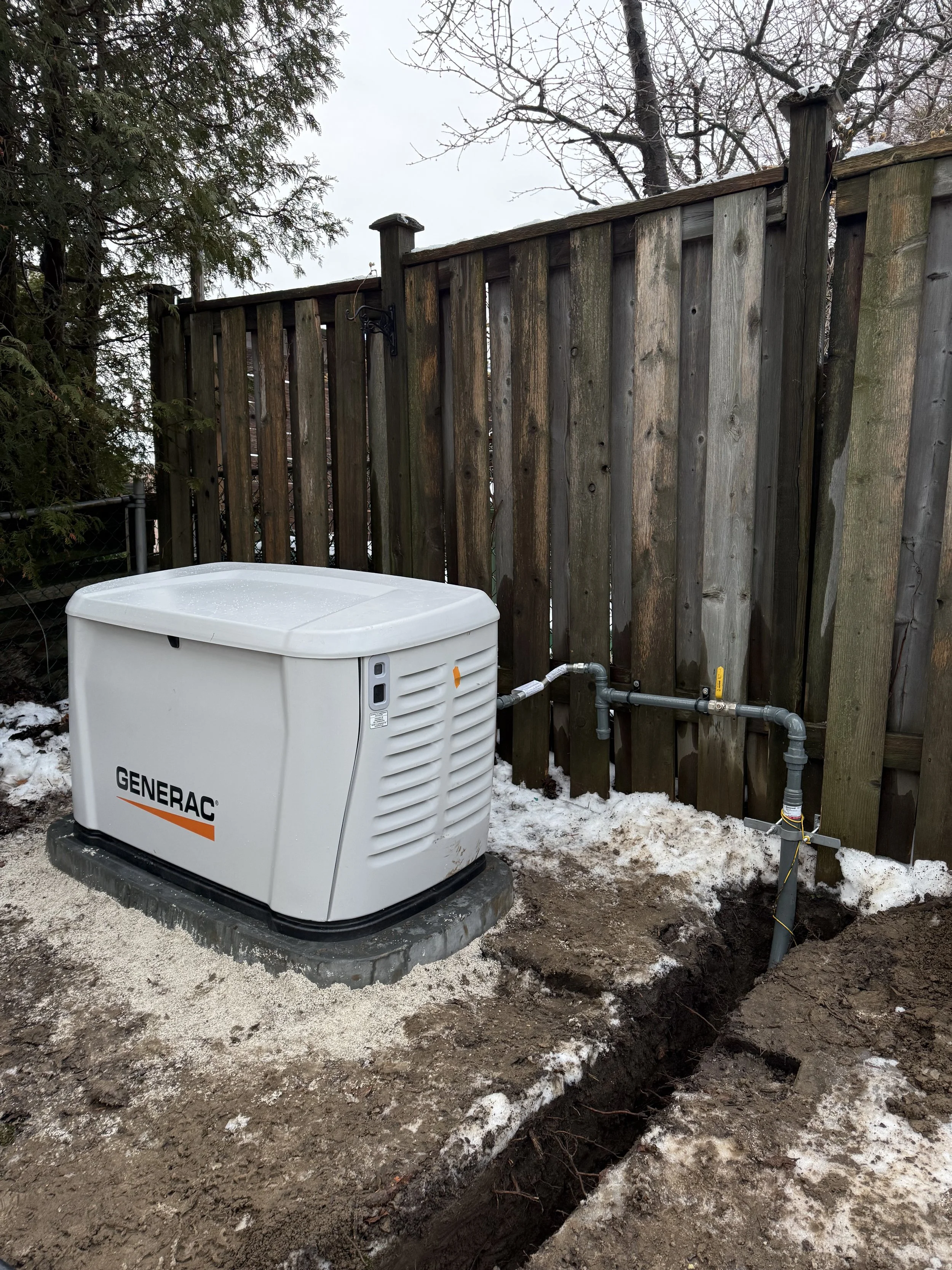 A Generac backup generator installed outdoors on a concrete pad next to a wooden fence, with exposed underground piping and snow on the ground.
