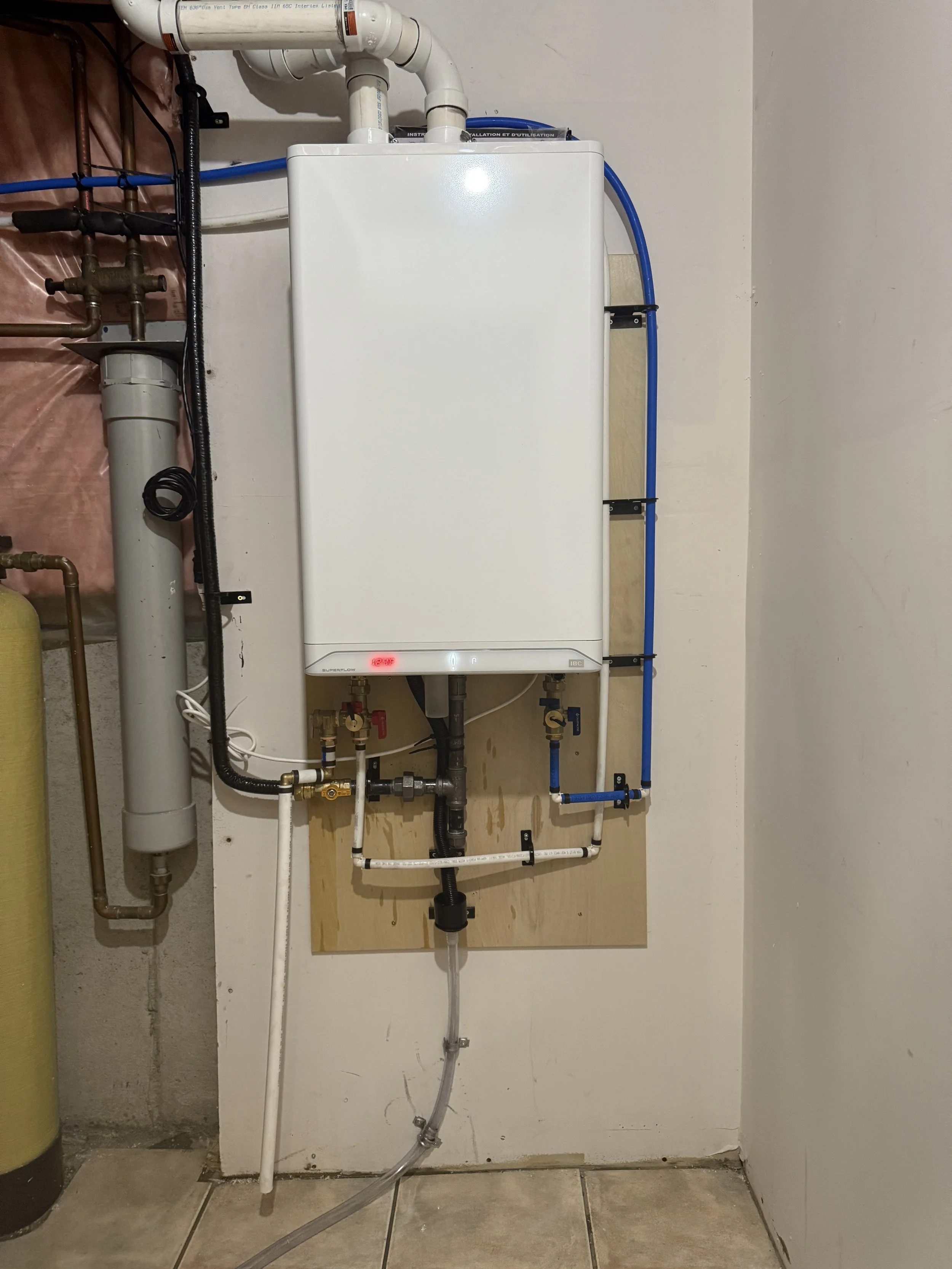 A white tankless water heater mounted on a wall with various plumbing pipes and valves connected to it, including white, black, and blue pipes. The heater has a red digital display light and is installed on a wooden board, with parts of the wall and 