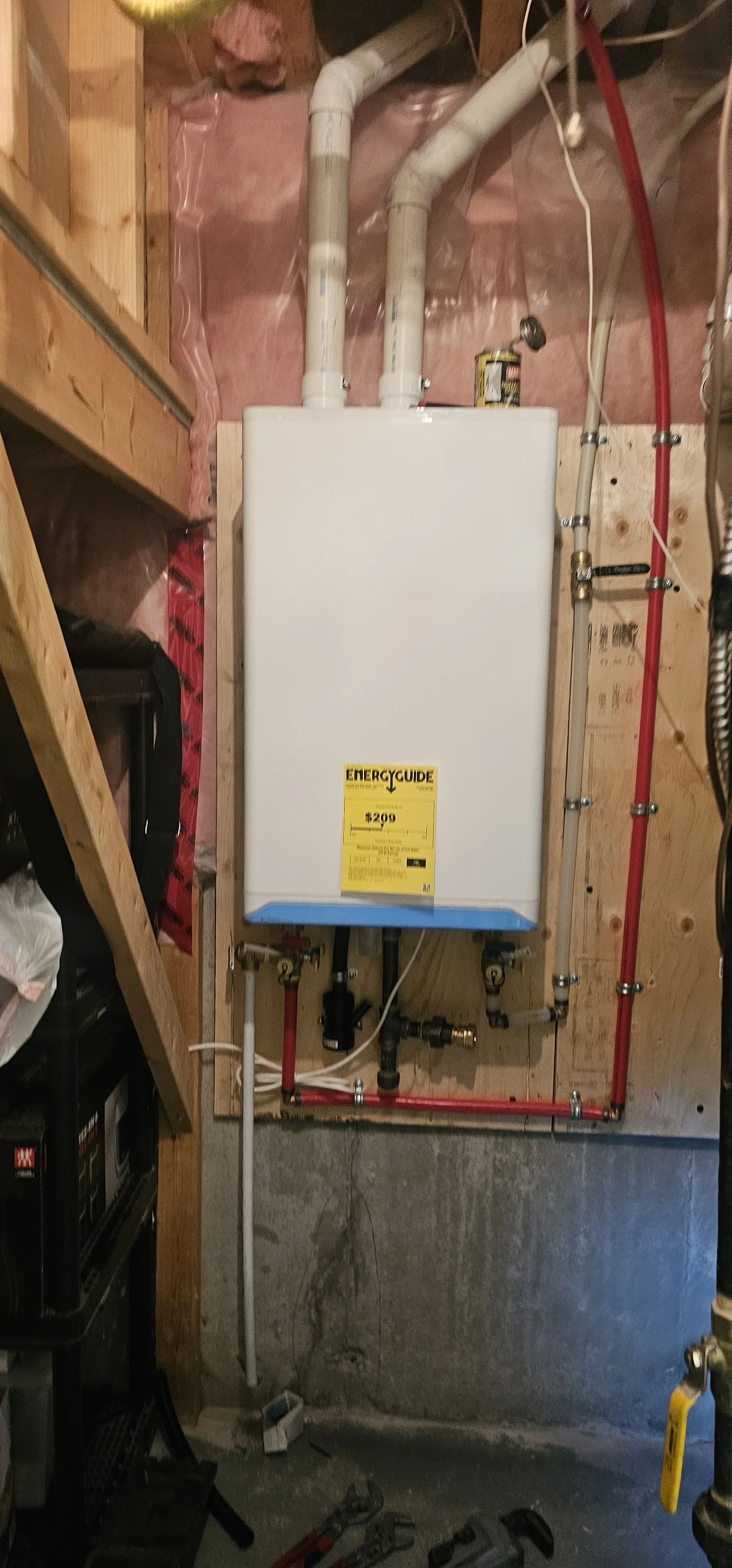 A tankless water heater installed on a wooden wall in a basement or utility room, with exposed plumbing pipes and an energy guide label.