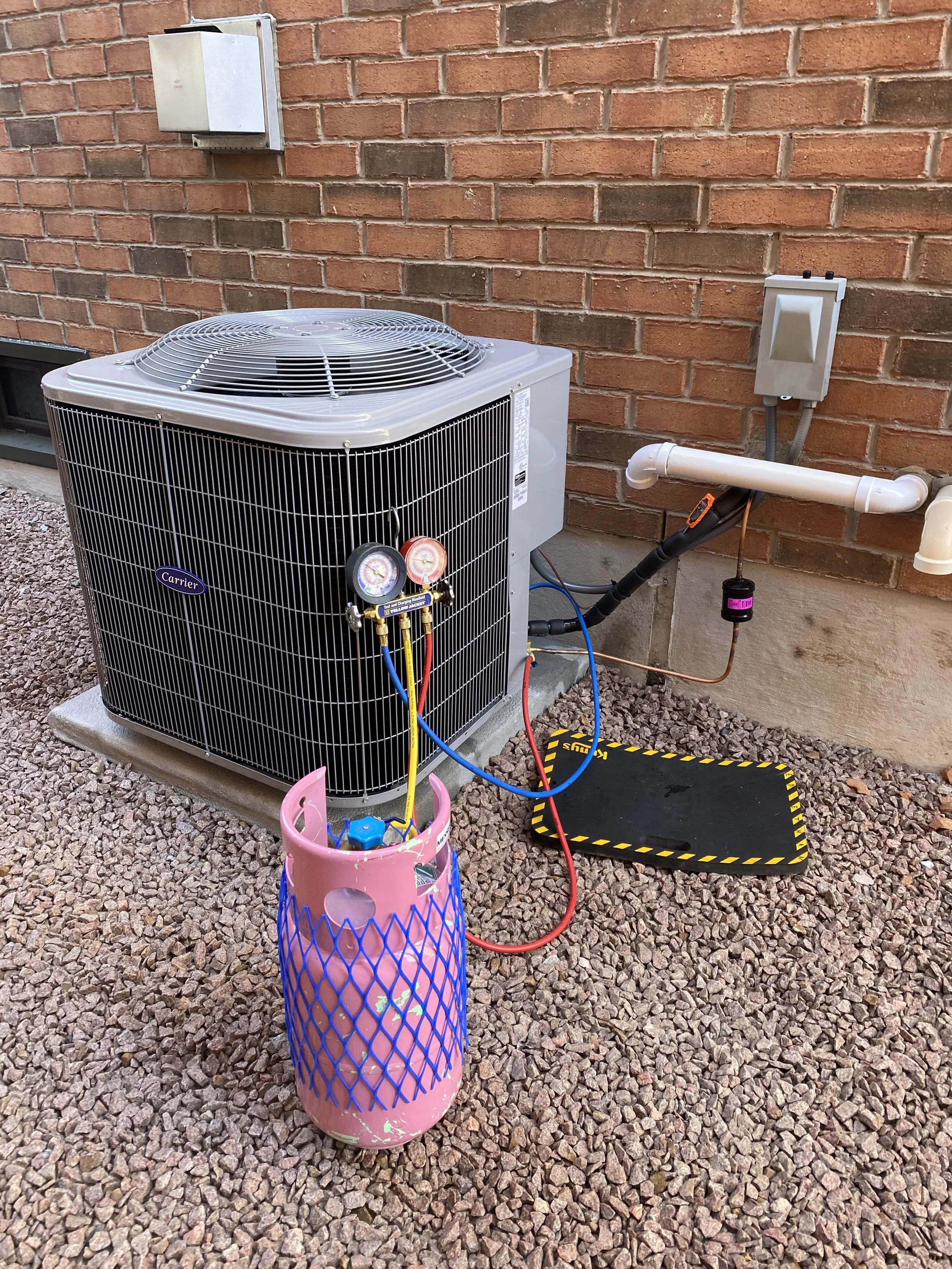 An outdoor HVAC unit connected to a pink refrigerant tank via hoses, situated on gravel next to a brick wall. The unit has gauges attached to the hoses, and various electrical components and piping are visible.