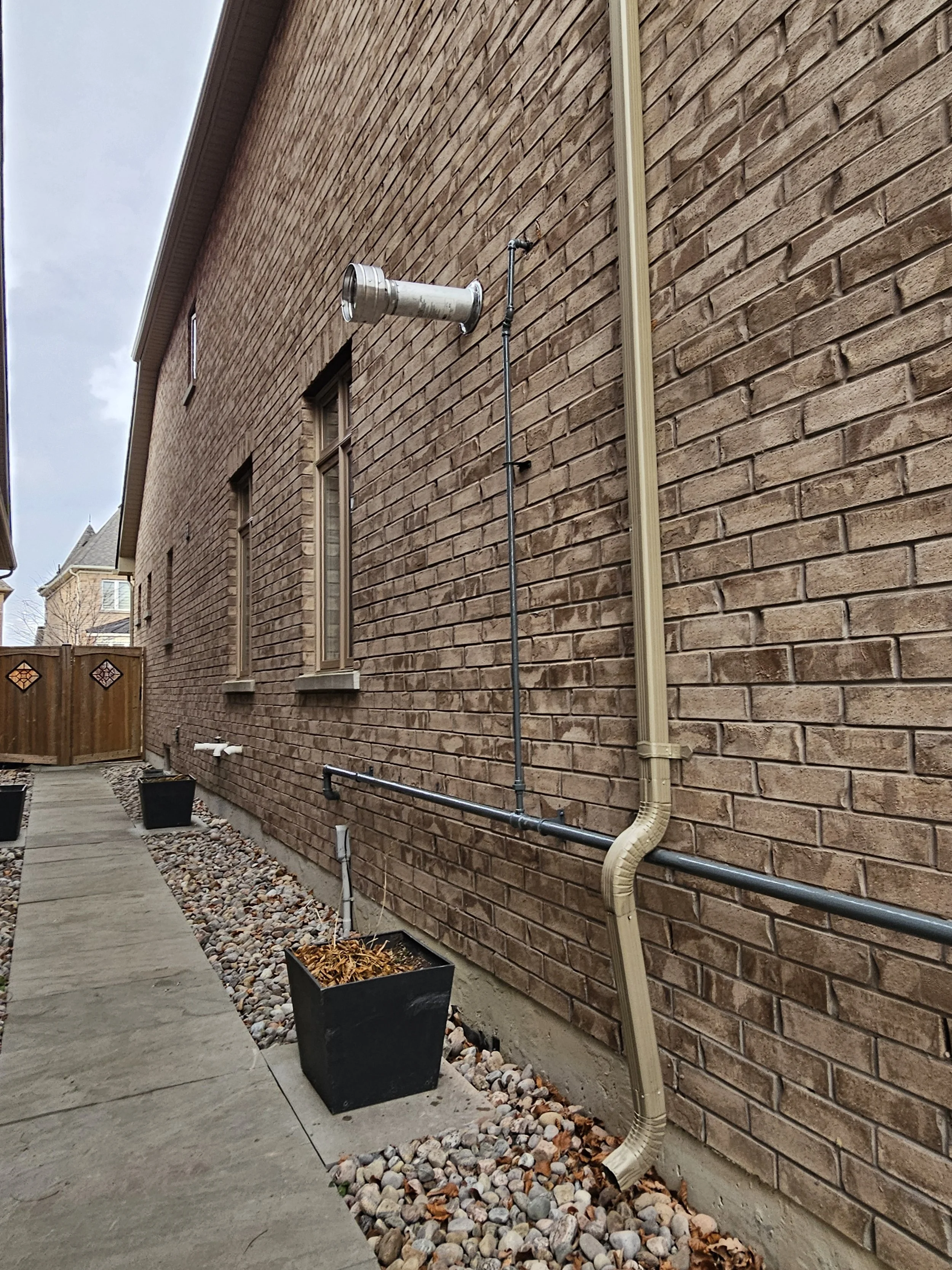 Side of a brick house with an outdoor exhaust pipe, electrical conduit, and a drainage pipe down the wall, a concrete sidewalk, and black planters with dried leaves.