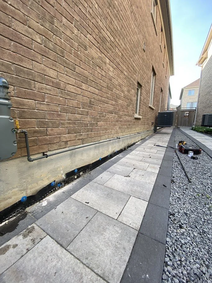Sidewalk construction along a brick building with new paving stones and partially completed pathway, construction tools and gravel nearby.