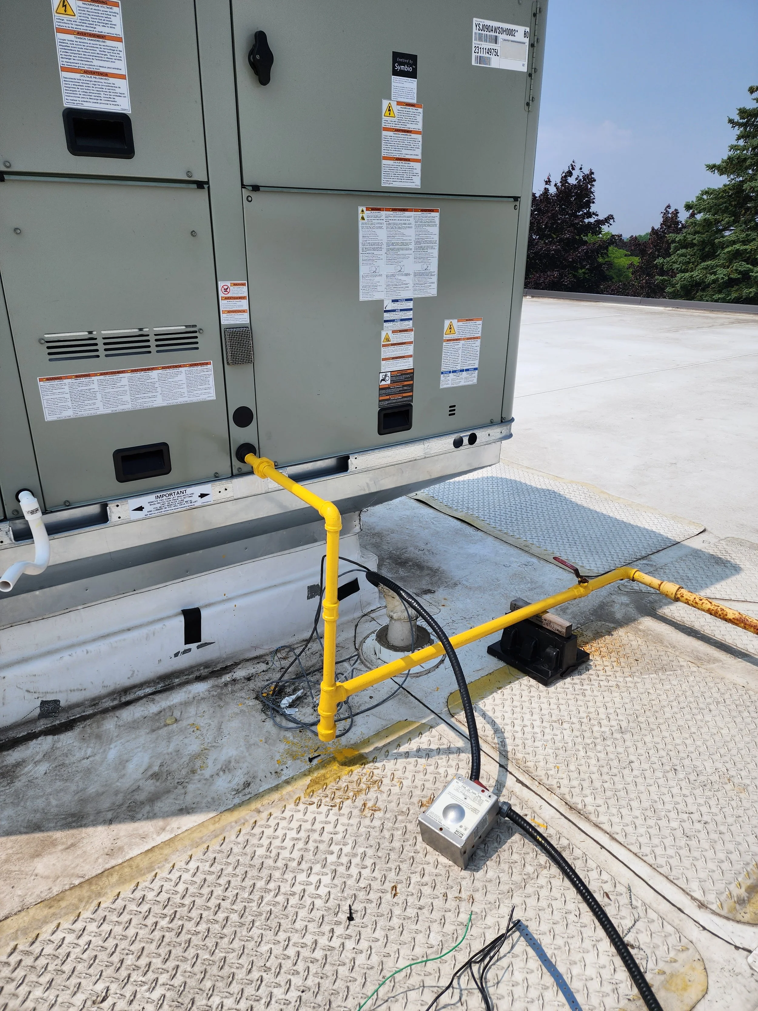 Close-up of commercial HVAC or electrical equipment on a rooftop with warning and instruction labels, connected by yellow and black pipes and wires.