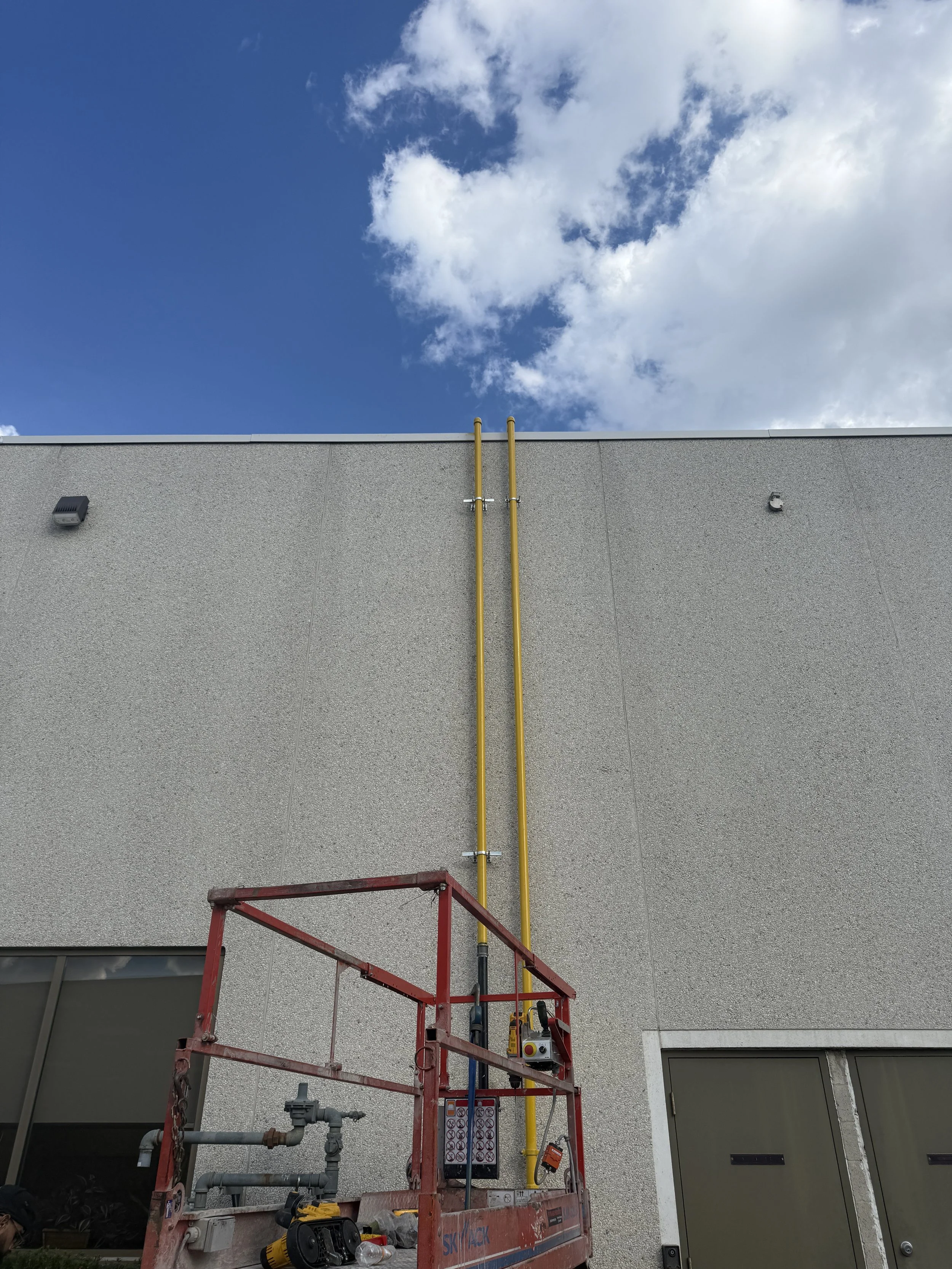 Exterior of a building with two yellow pipes running vertically up the wall. There is a red scaffolding platform in the foreground with tools and equipment.