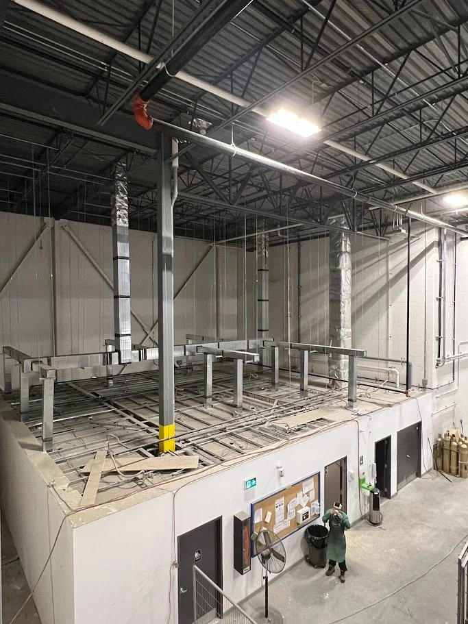 Industrial space under construction with elevated platform, pipes, and ventilation ducts, a worker wearing protective gear, and construction materials.