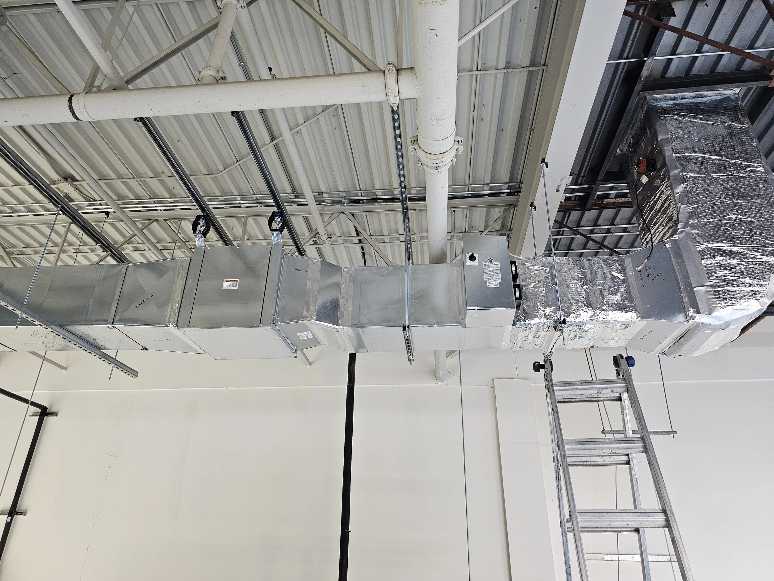 Industrial air duct system hanging from ceiling, with aluminum insulation and a ladder nearby in a warehouse or commercial building.