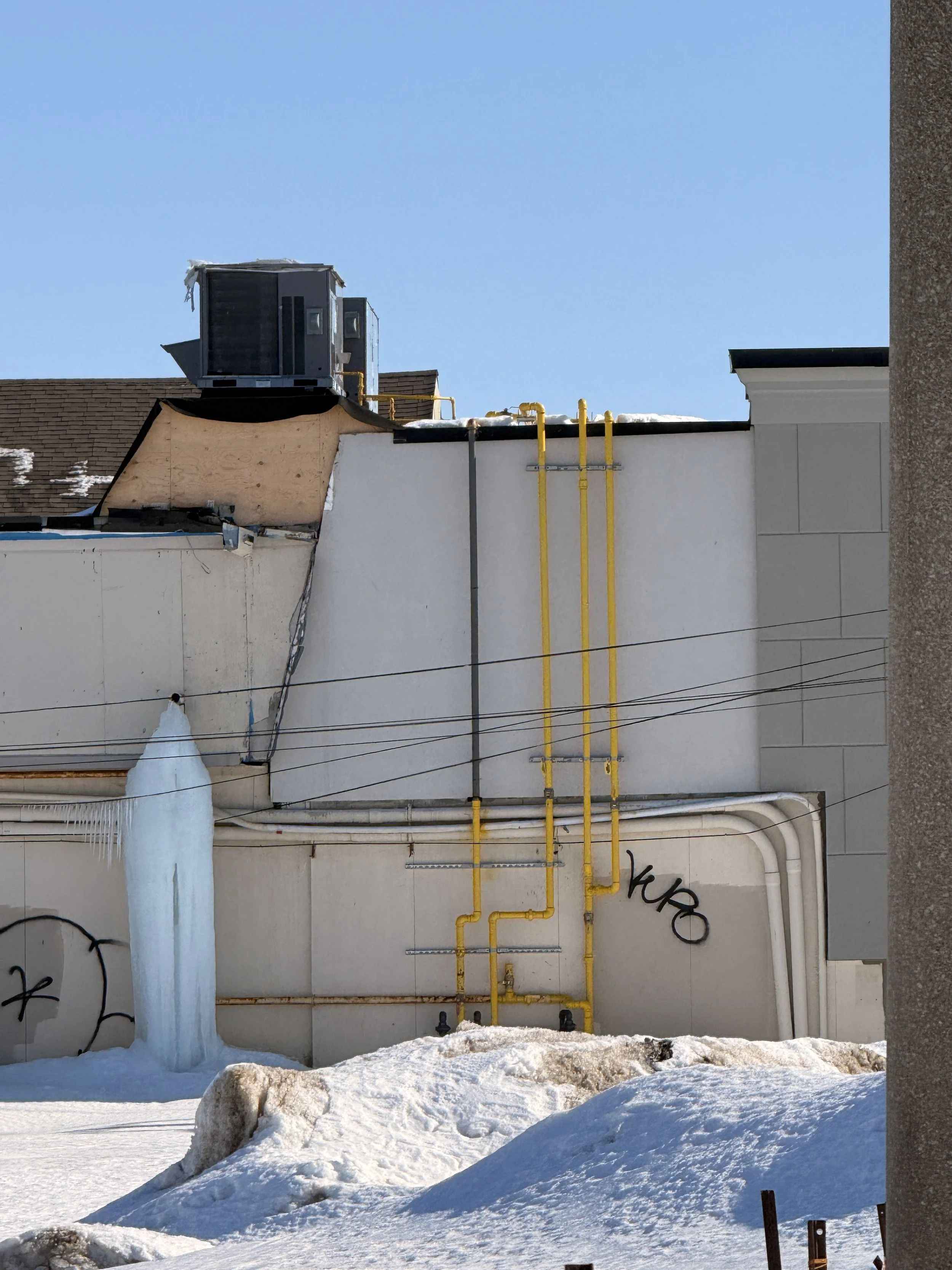 Building rooftop with yellow gas pipes, an air conditioning unit, and icicles hanging from the roof edge, snow on the ground and a clear blue sky.