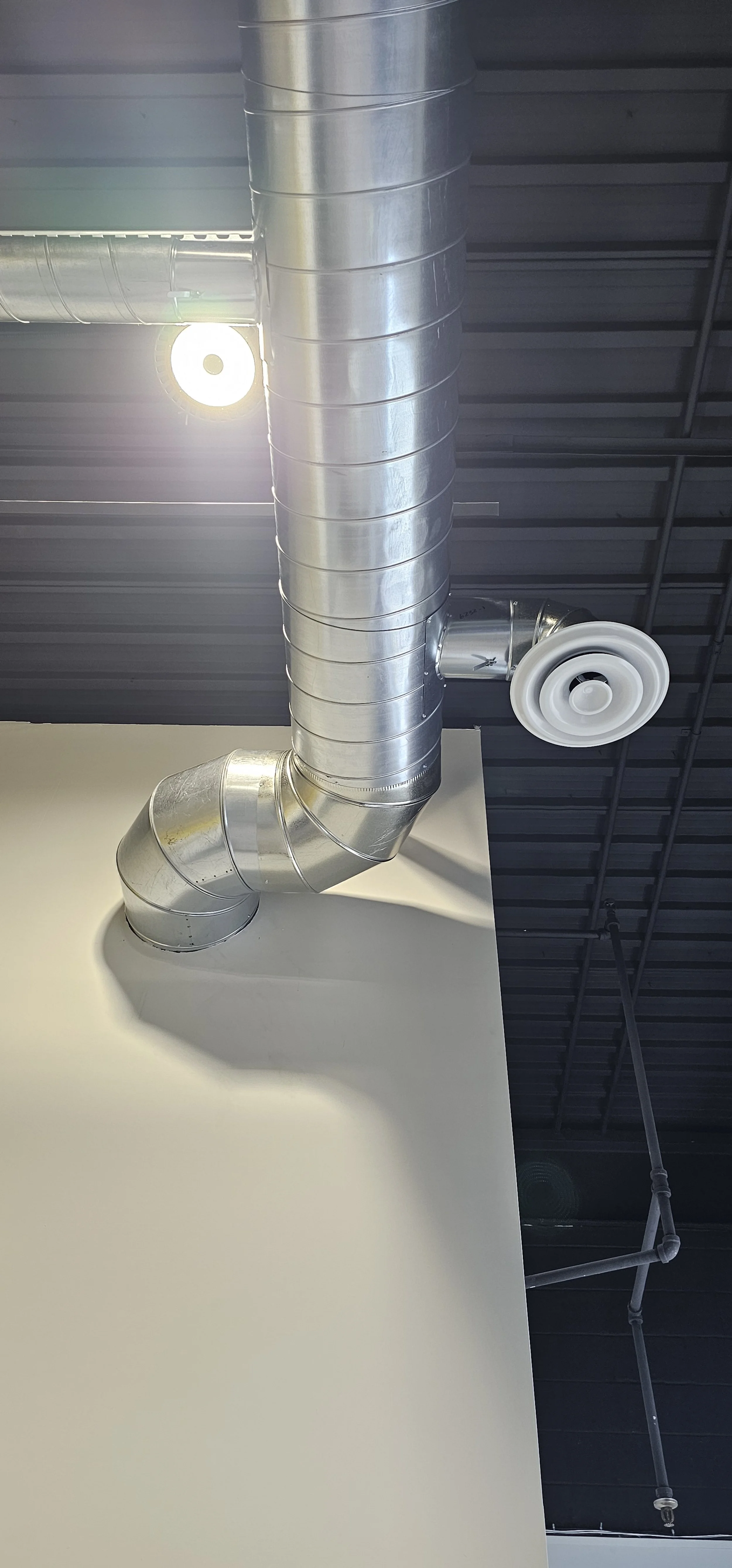 Industrial ceiling with metal ventilation ductwork and a white circular ceiling vent, with a black ceiling and pipes visible in the background.
