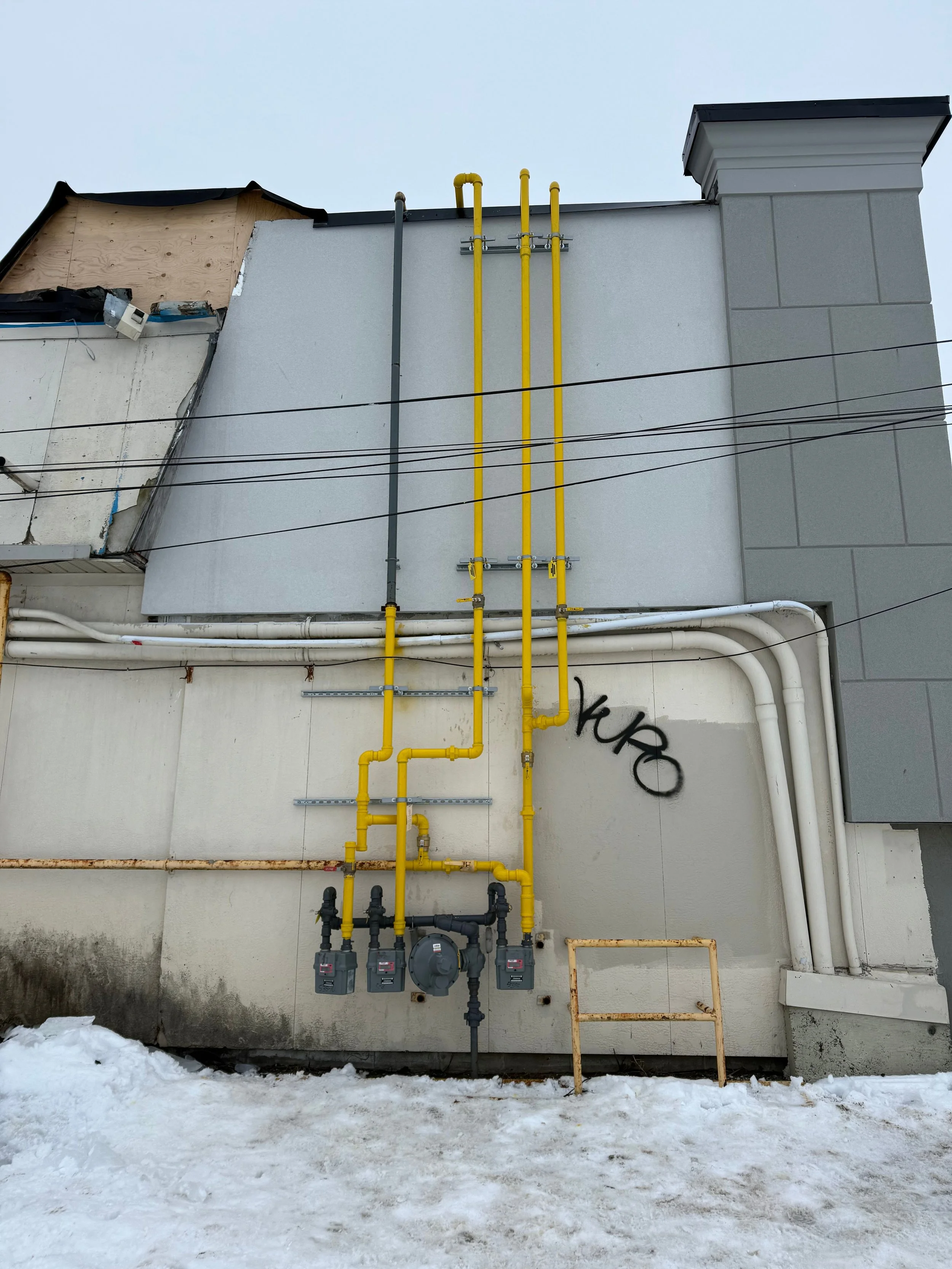 Exterior wall with yellow gas pipes, white and gray electrical conduits, and graffiti on the wall, with snow on the ground.