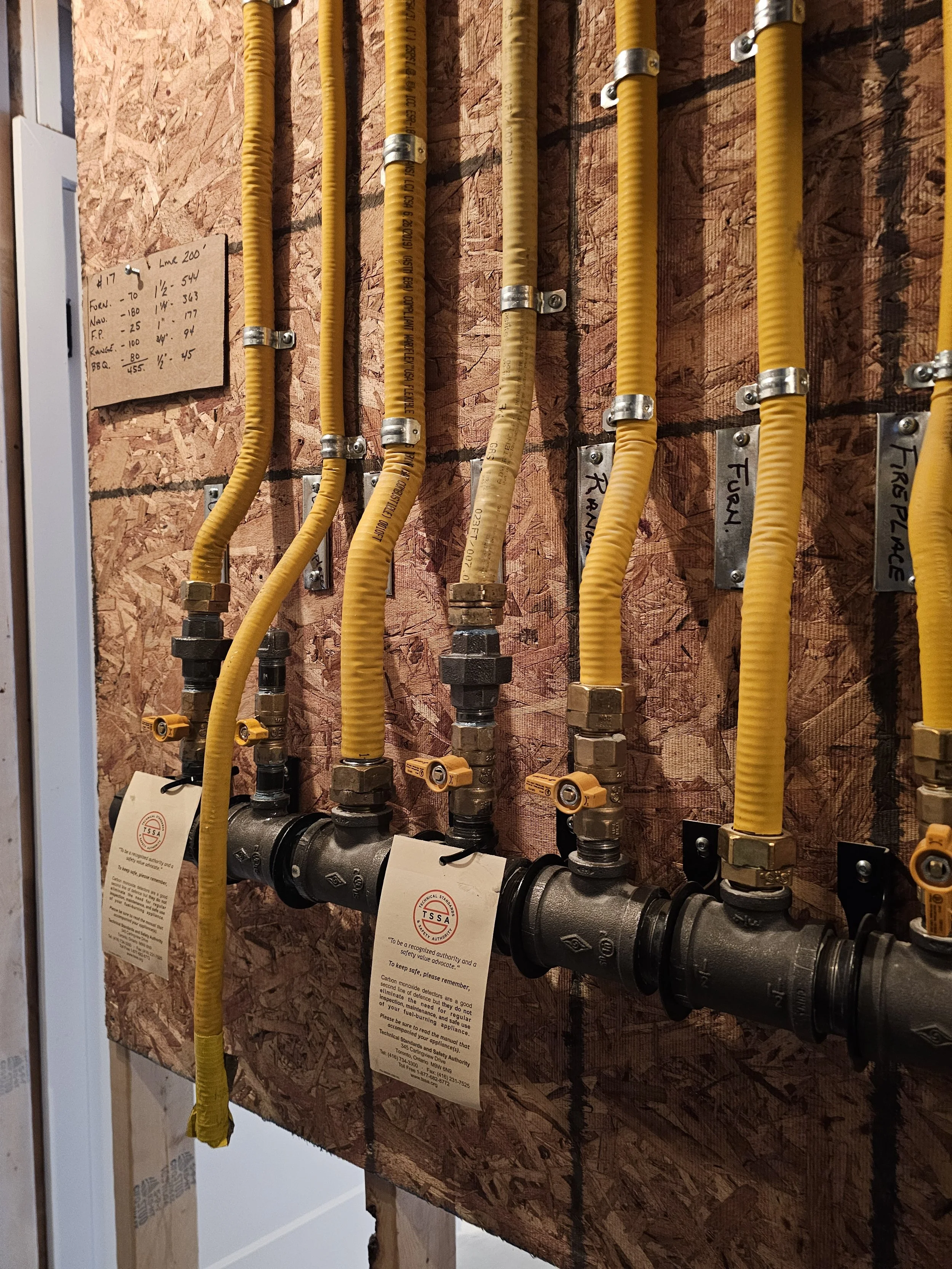 Yellow flexible pipes connected to a black metal pipe and managed with metal clamps on a wooden wall, with handwritten labels and tags attached.