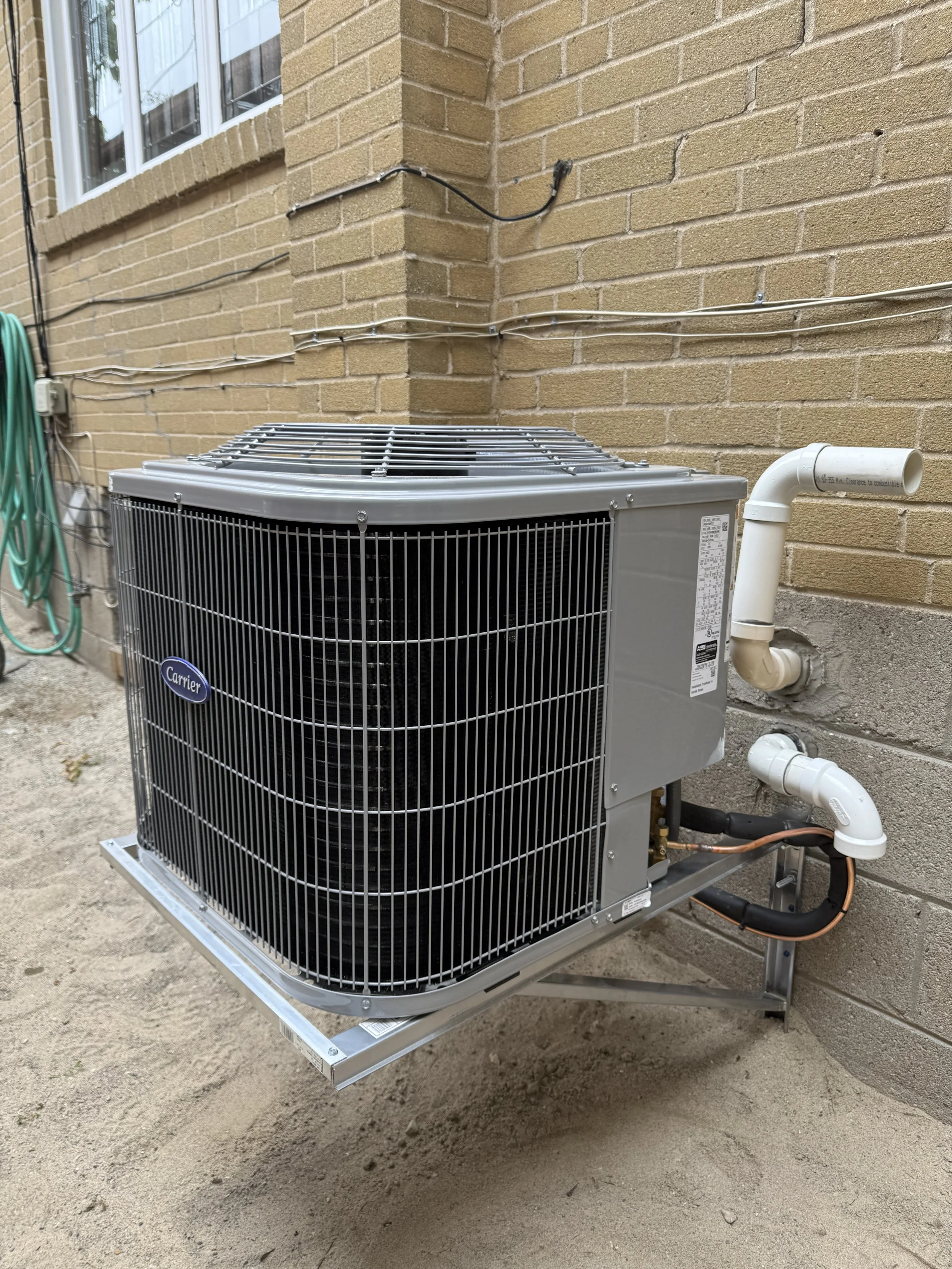 An outdoor HVAC unit installed next to a brick house wall, with pipes and a green garden hose hanging on a hook nearby.