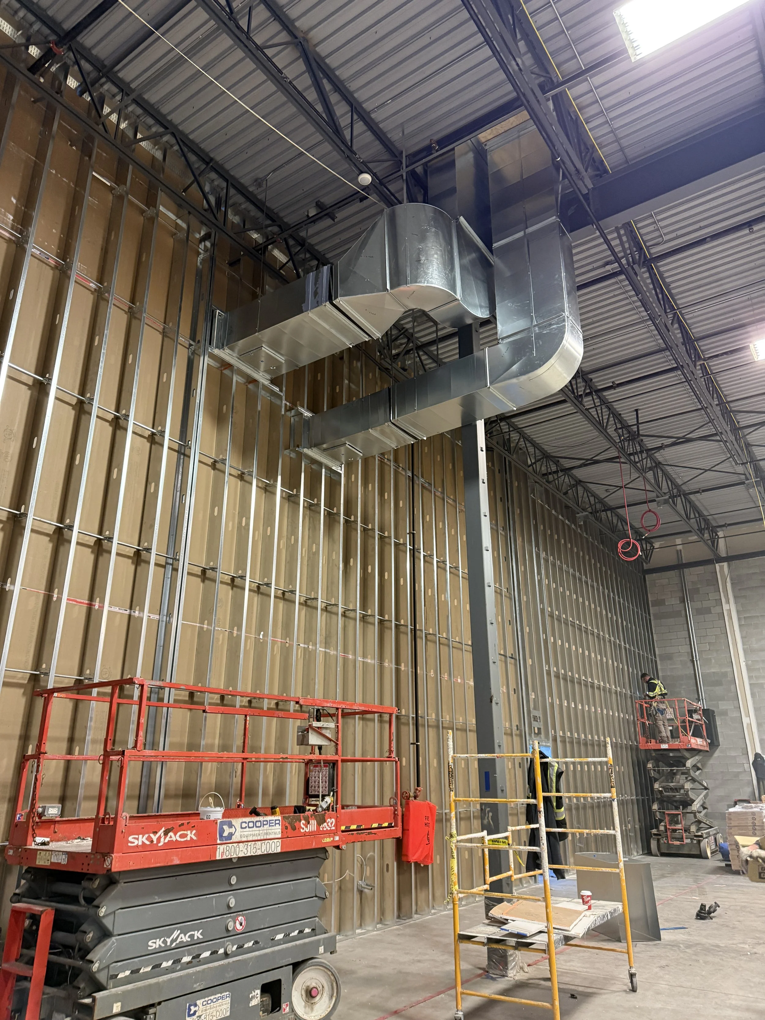 Construction workers installing ductwork on the ceiling of a large industrial building with scaffolding and construction equipment.