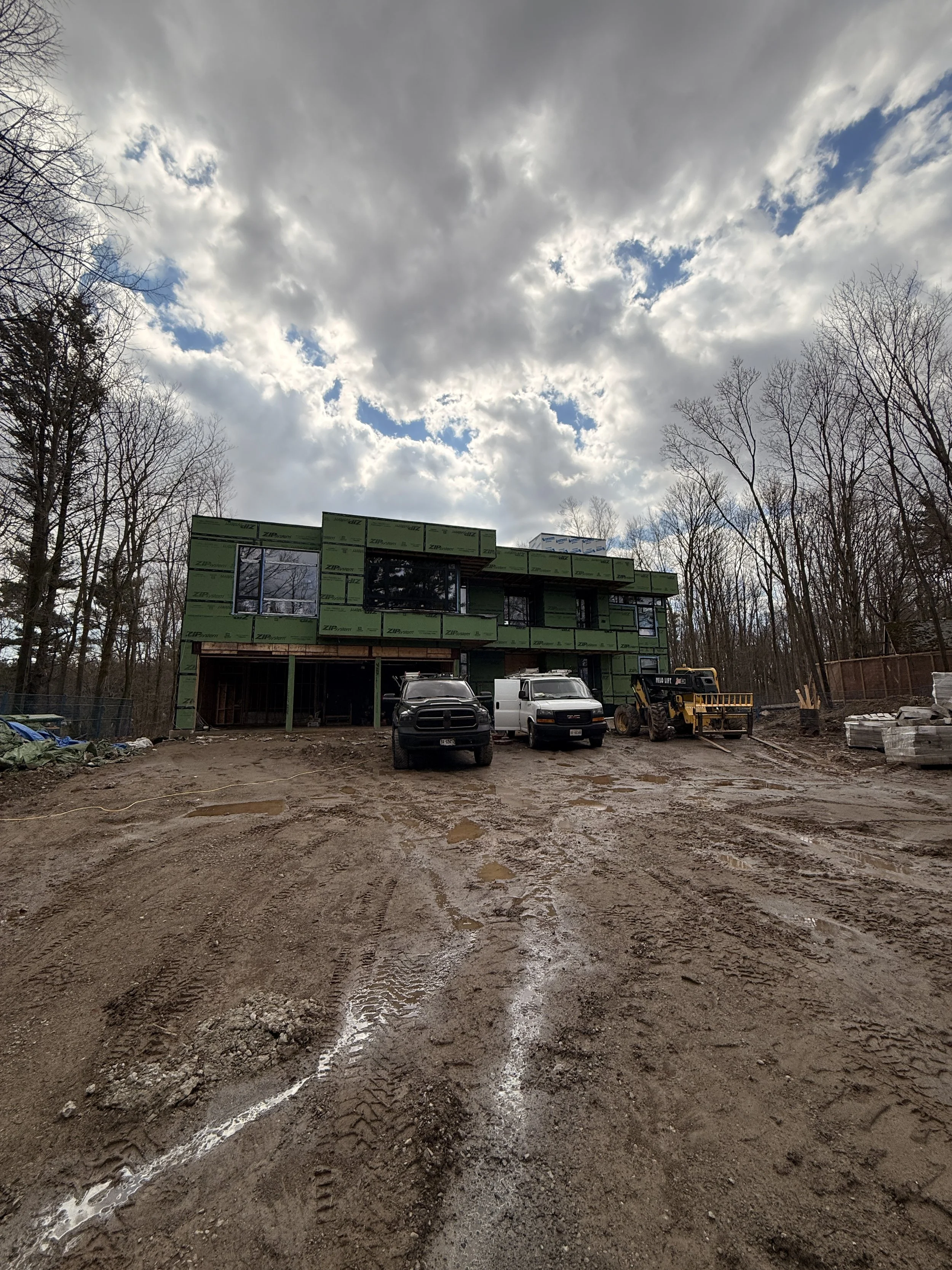 Construction site with a partially built house covered in green sheathing, three vehicles parked in front, mud and tire marks in the foreground, and cloudy sky overhead.