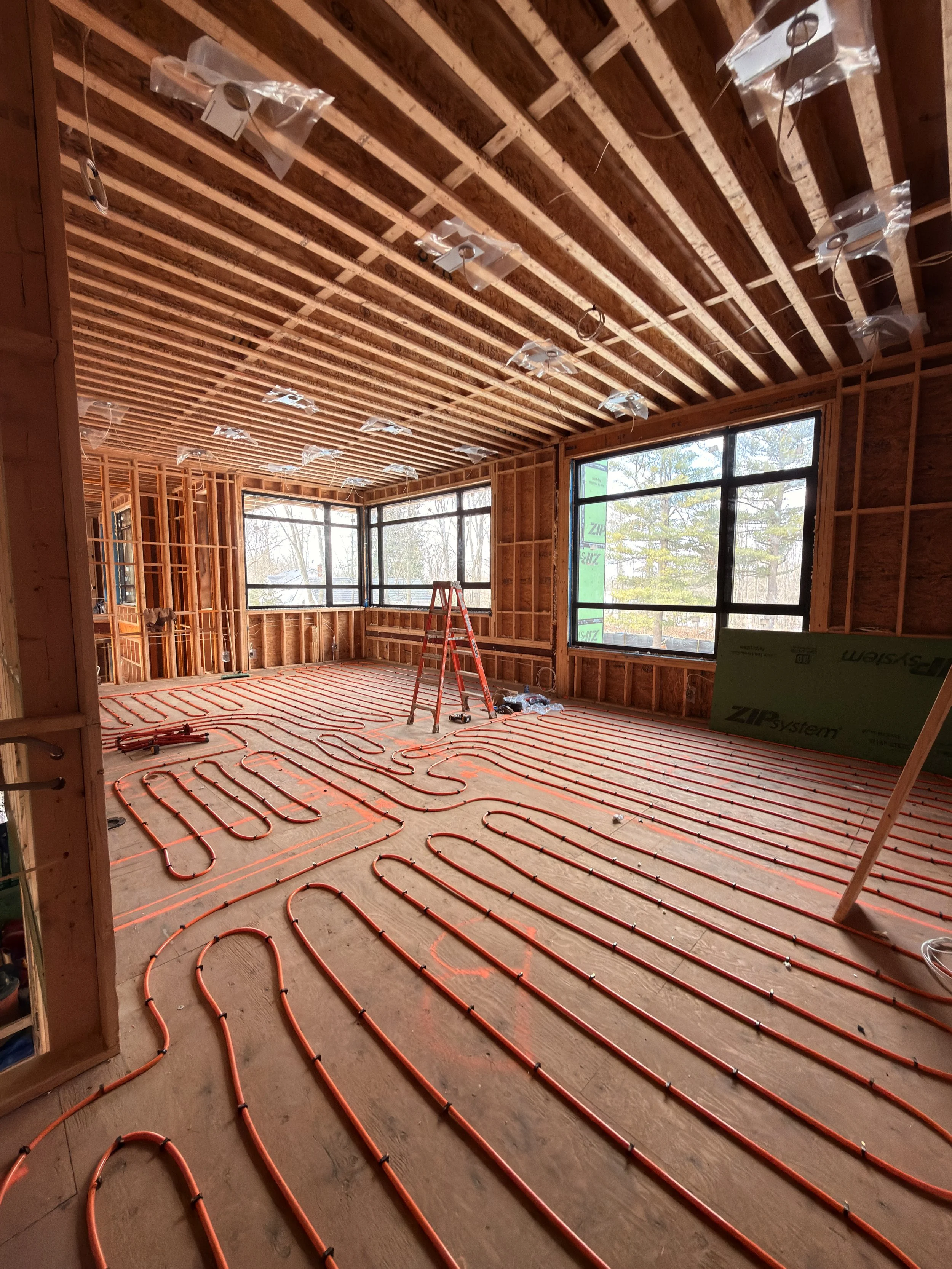 Interior of a house under construction with exposed wooden framing, orange radiant floor heating pipes installed on the floor, and large windows providing natural light.