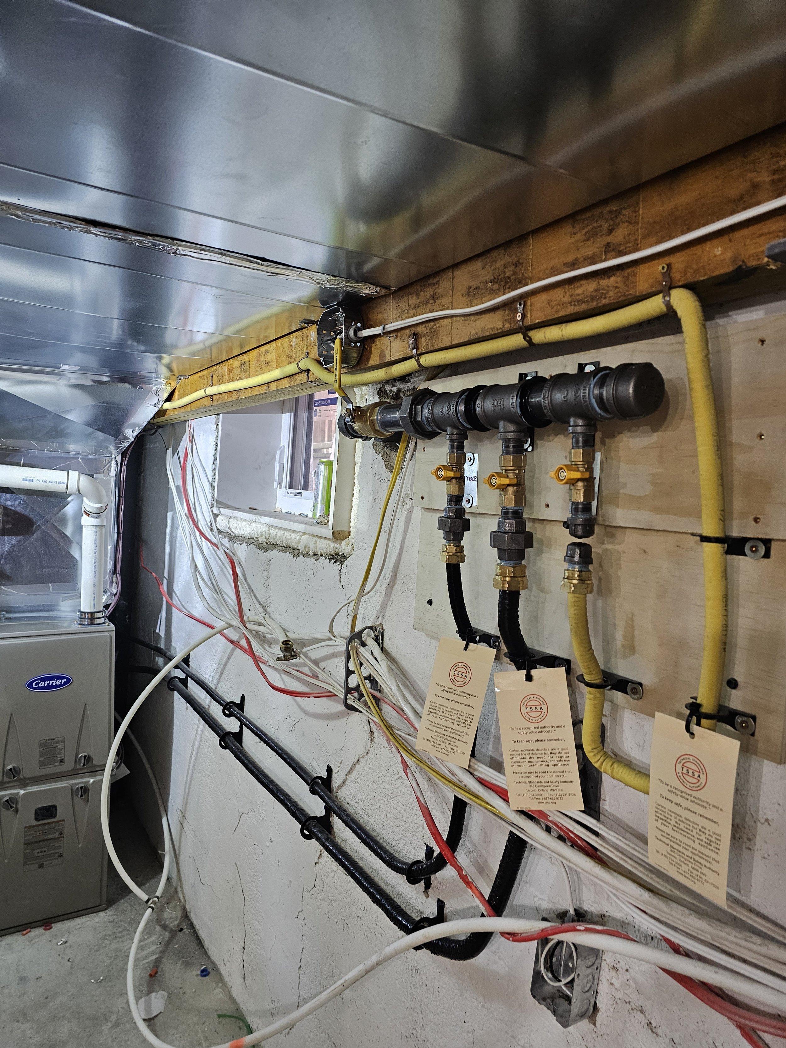 Plumbing and electrical setup in a basement or utility room, with yellow hoses, black pipes, and electrical wiring installed on a wooden wall next to a small window.
