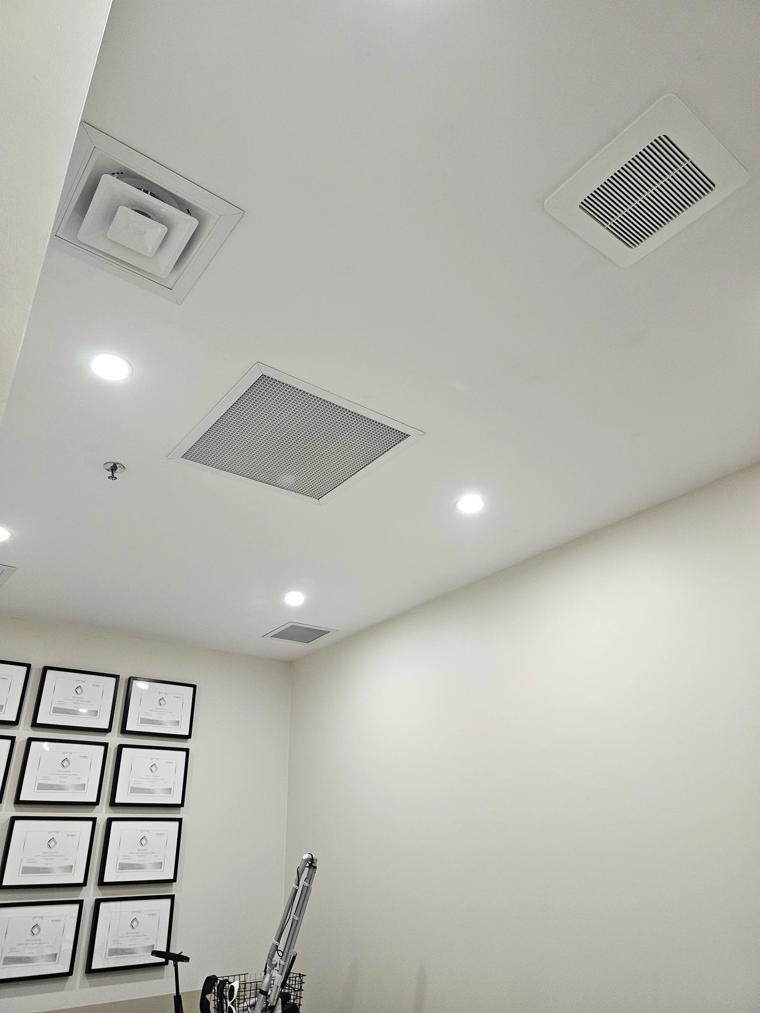 Ceiling with multiple air vents and recessed lights in a room with framed certificates on the wall.