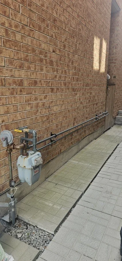 Close-up of a brick wall with utility gas meters and pipes attached near the ground, along with a section of paved walkway and a wooden fence in the background.