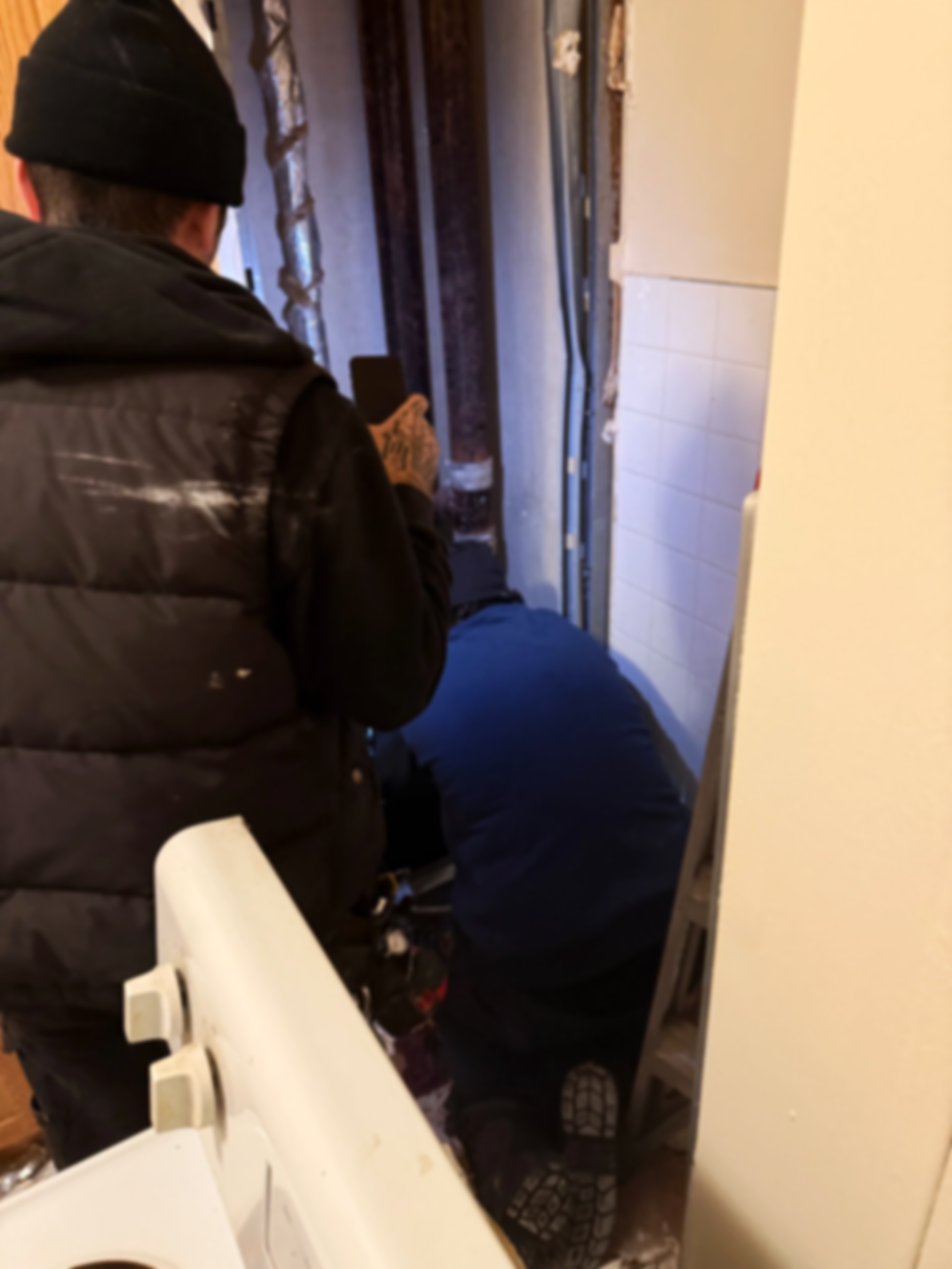 Two men working on a plumbing or renovation project inside a small room, one standing and the other kneeling near the floor.