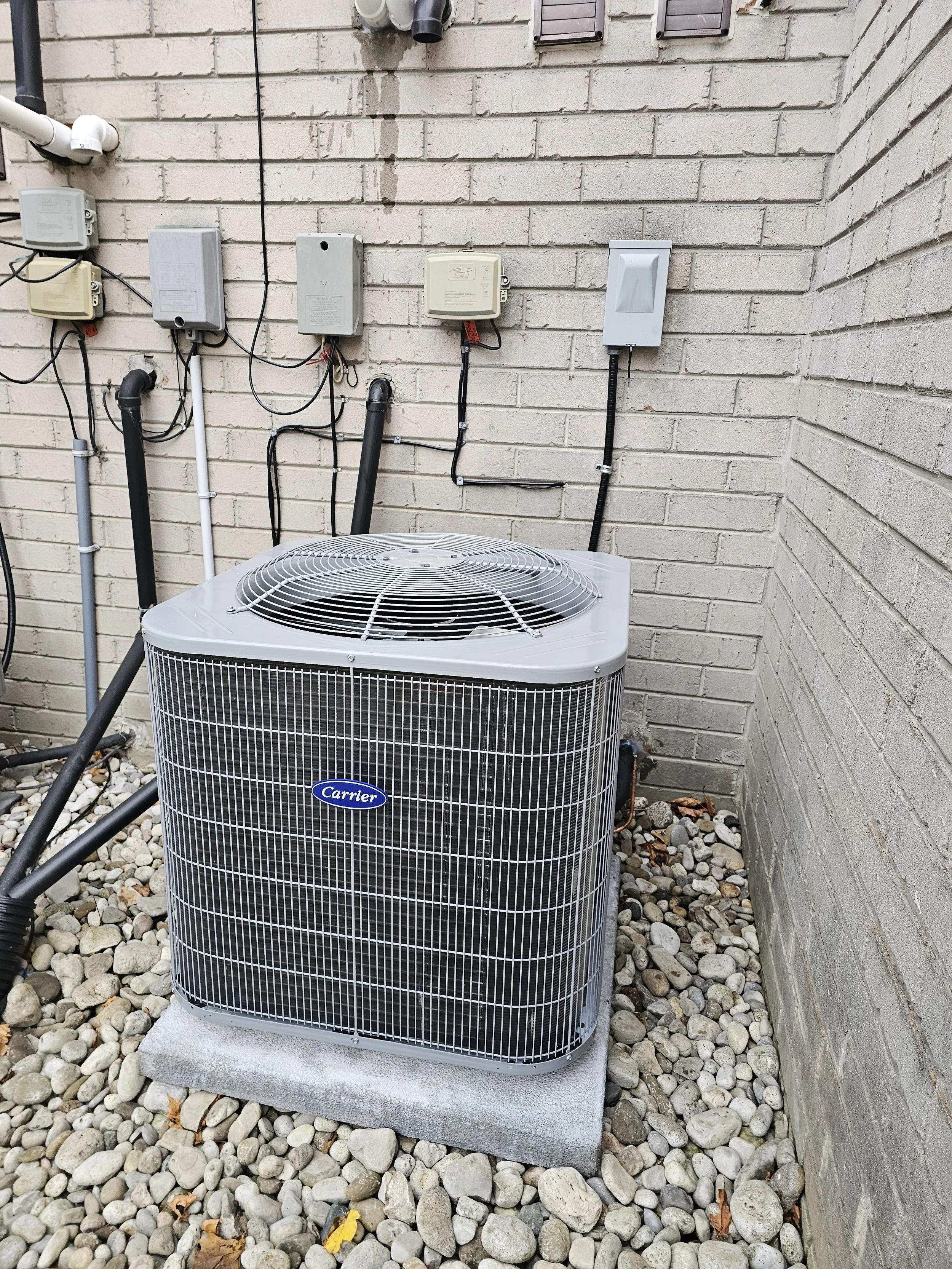 An outdoor air conditioning unit by Carrier is installed on a concrete pad near a brick wall, with several electrical boxes and pipes mounted on the wall above it, and small rocks scattered on the ground.