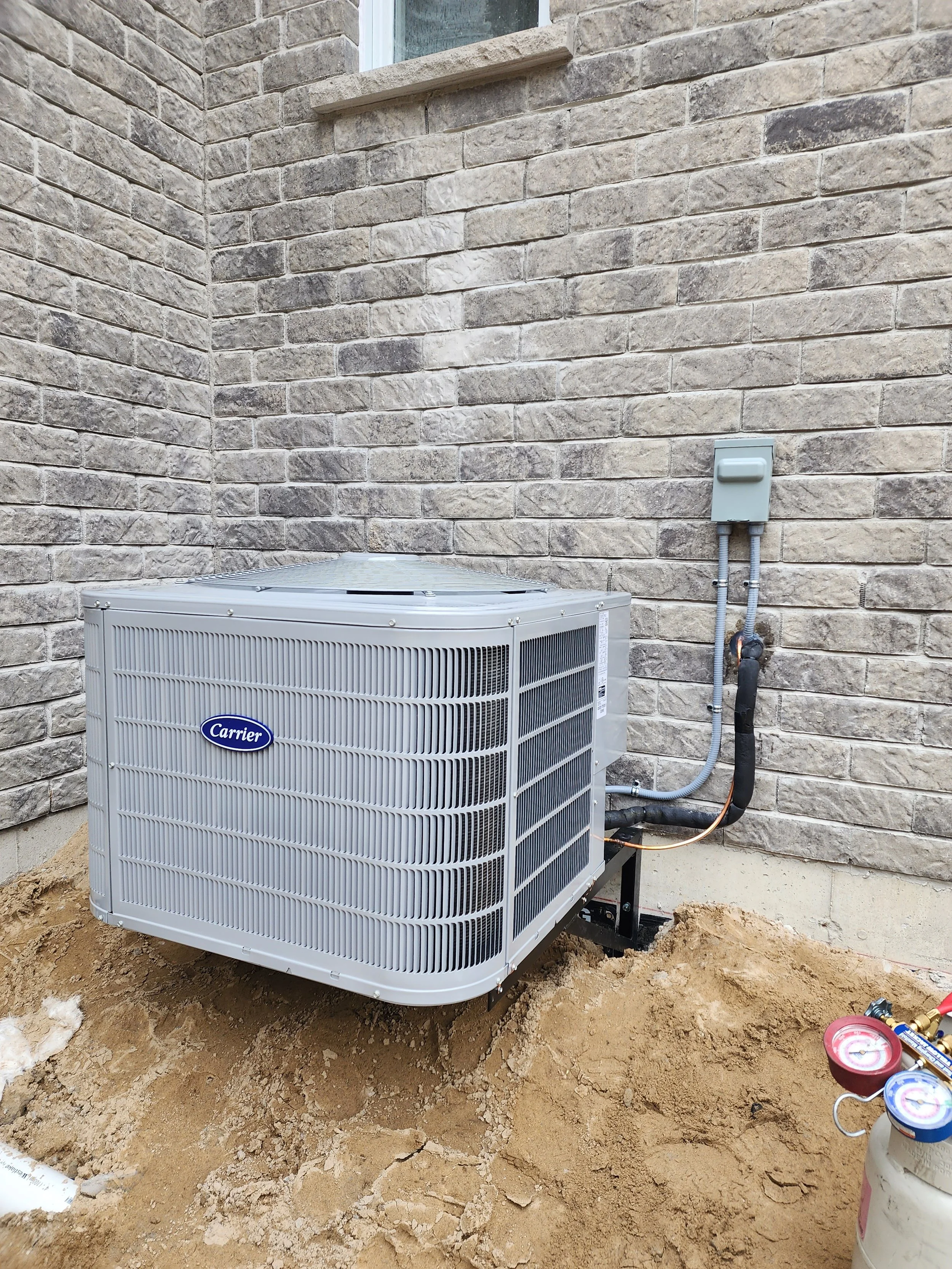 An outdoor air conditioning unit installed next to a brick house wall, with electrical wiring and pipes, and ground work around it.