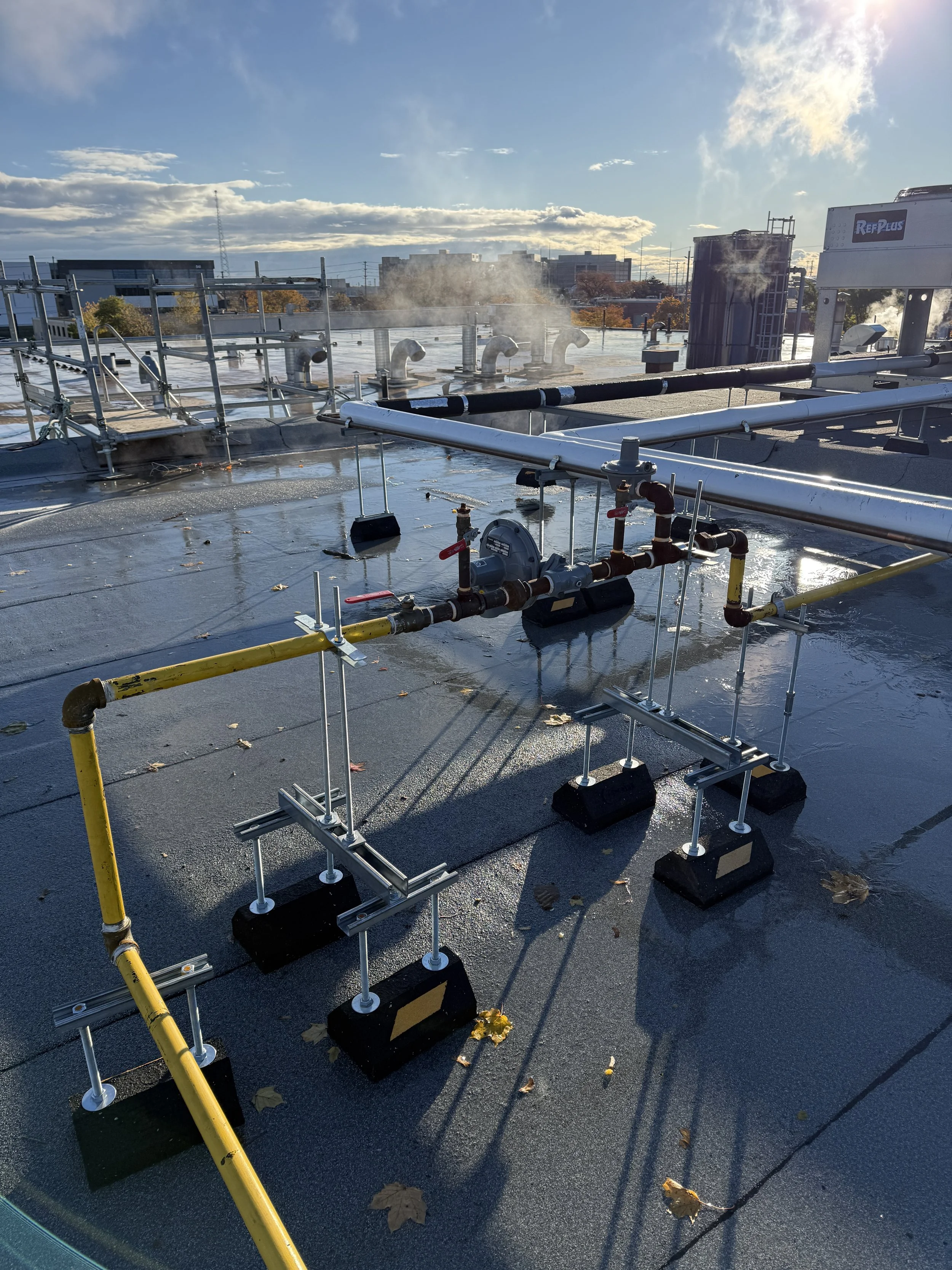 On a rooftop, various pipes, valves, and equipment related to HVAC or building systems, with some steam or vapor emissions, under a partly cloudy sky during daytime.