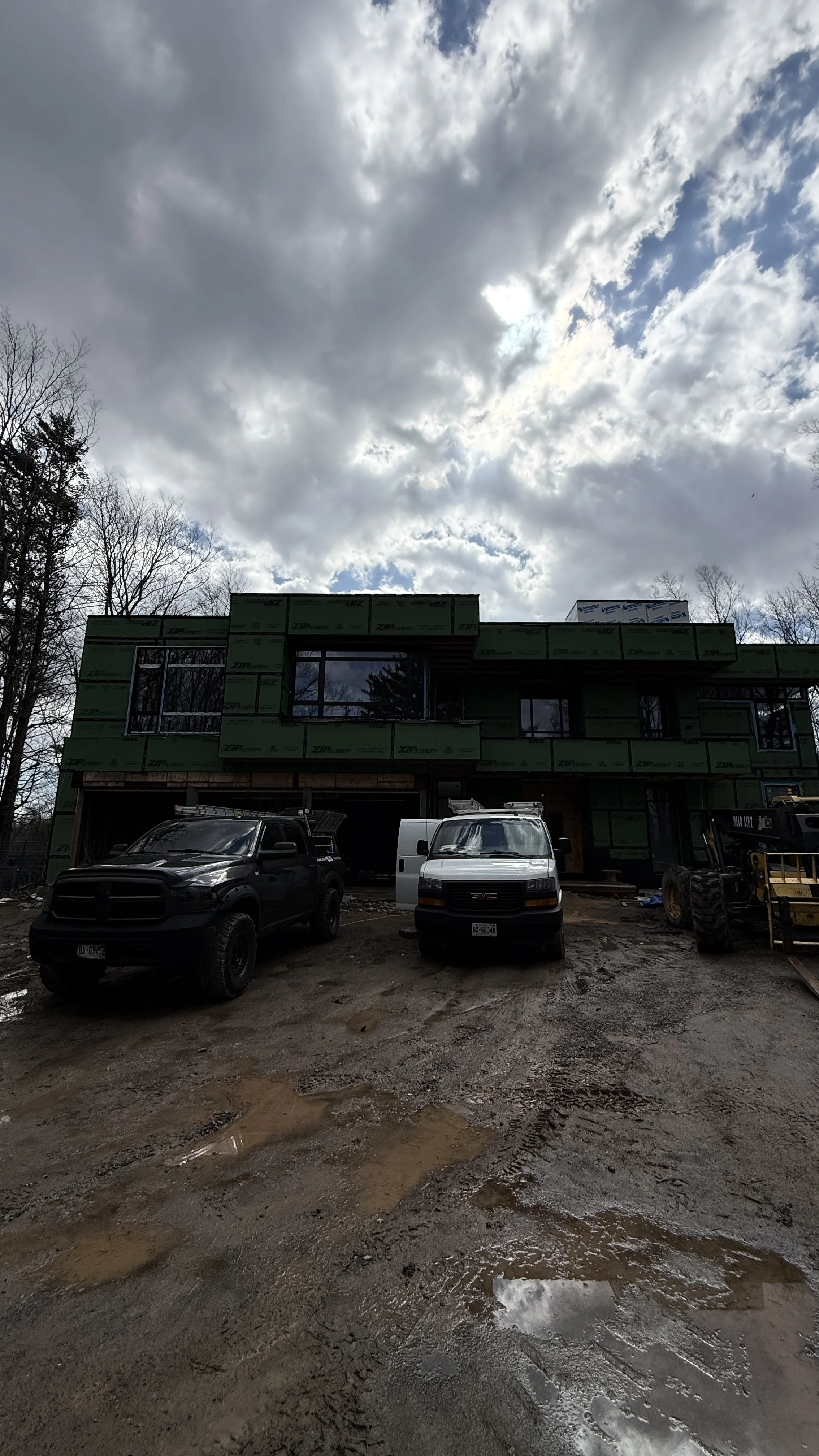 Construction site with three vehicles parked in front of a partially built house under cloudy sky.