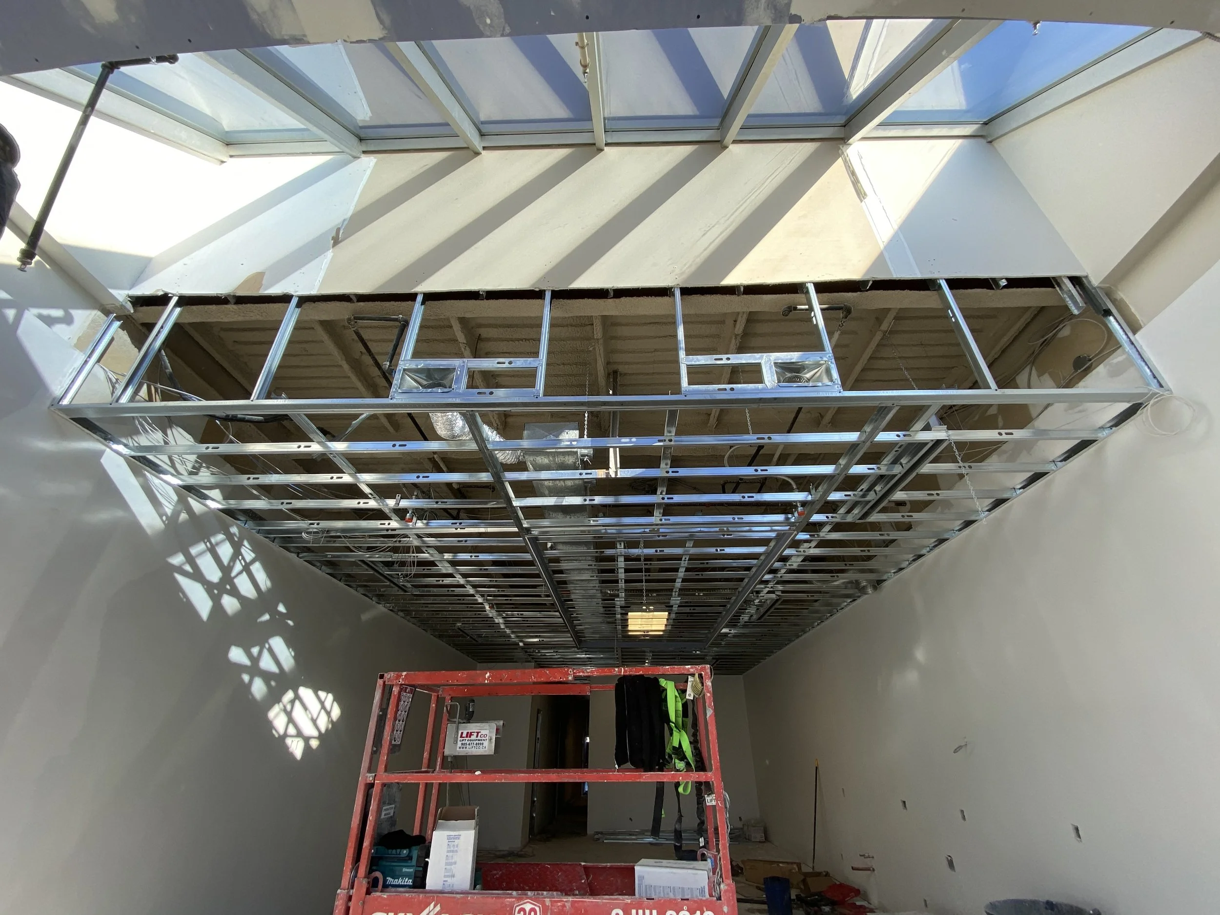 Construction site with metal framing for a ceiling or wall, a red lift with tools and gear, and a large window letting in sunlight.