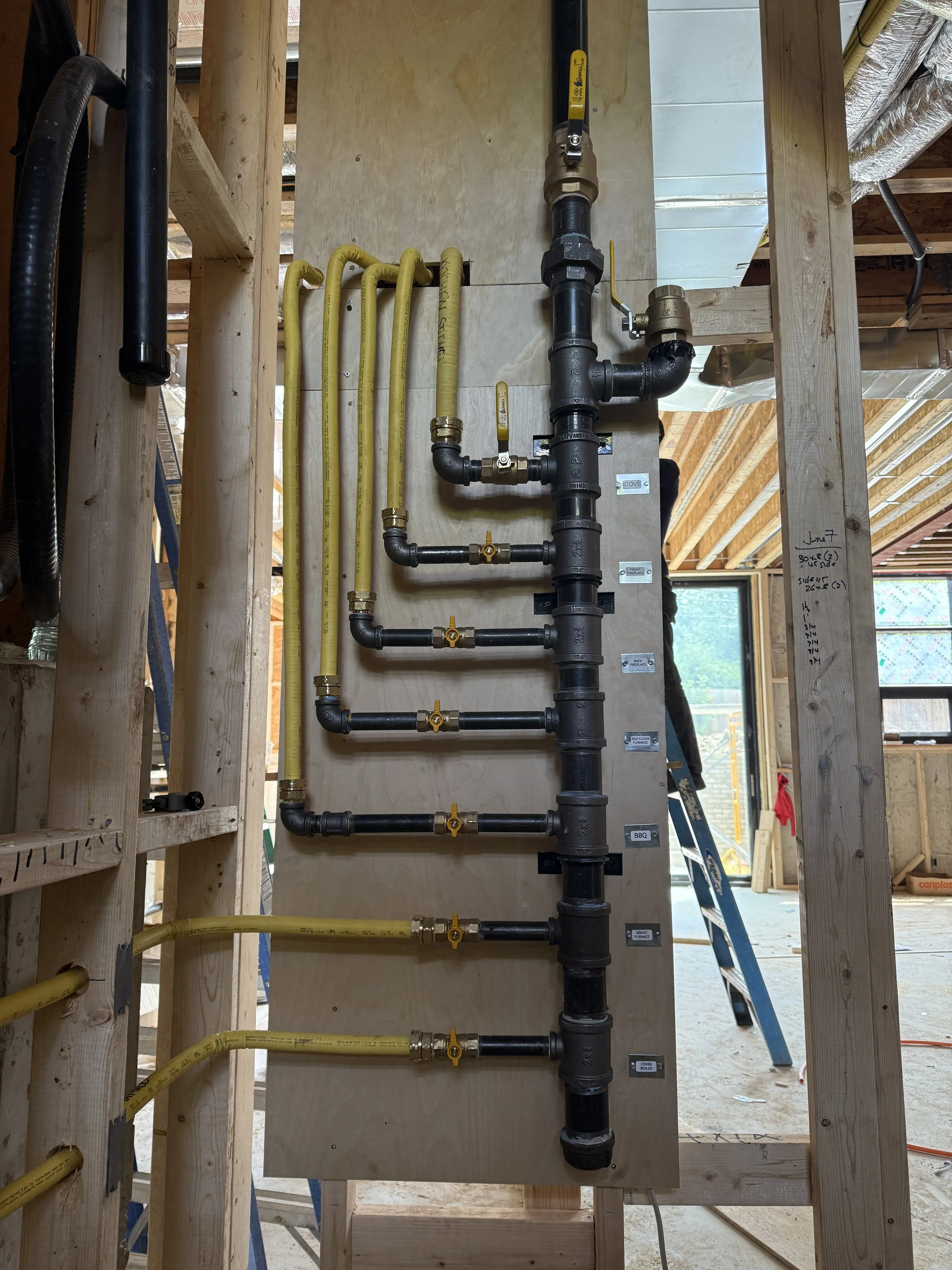 Construction site with plumbing pipes and yellow gas lines mounted on a wooden board, with various valves and fittings, and framing for walls in the background.