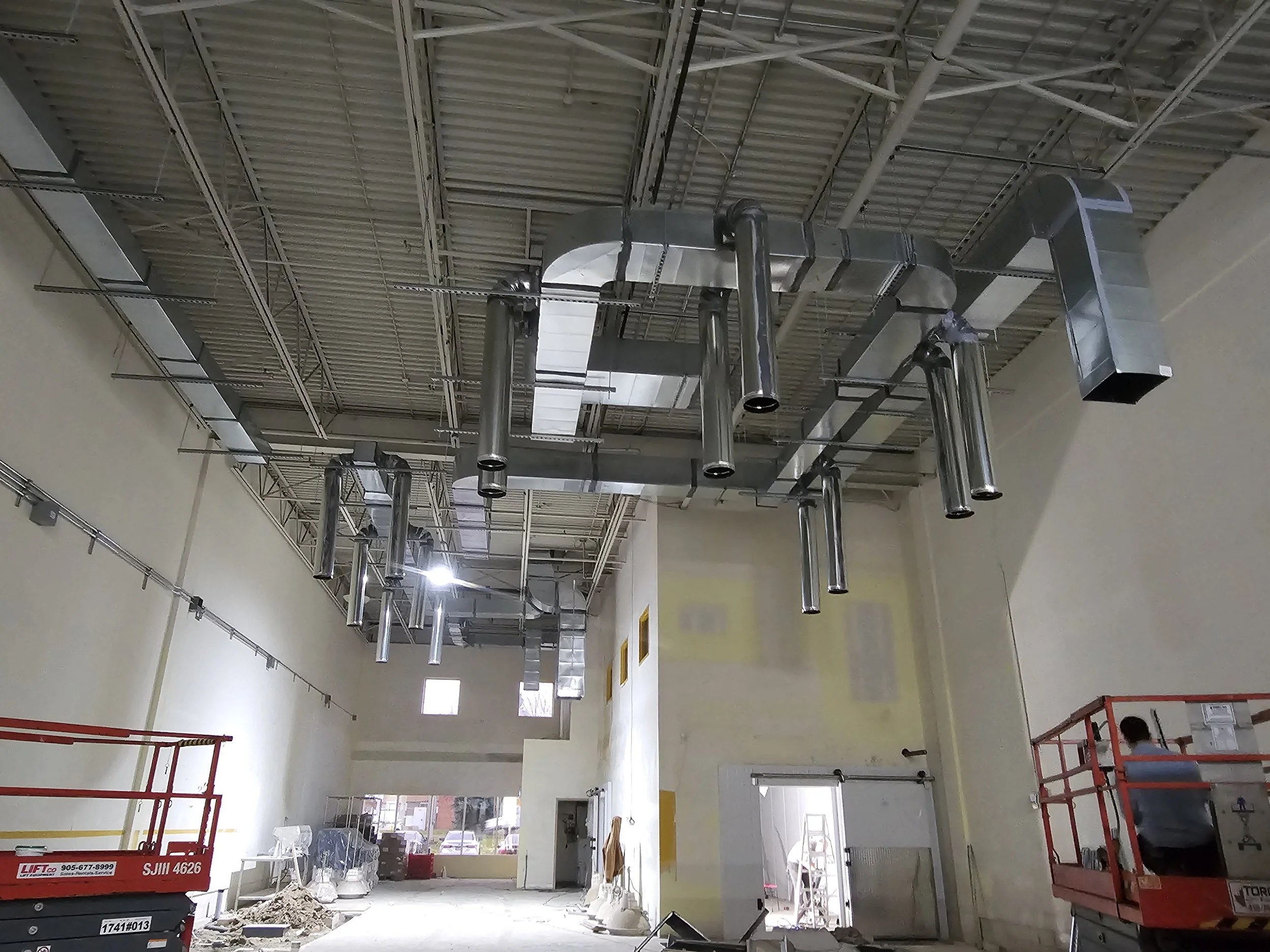 Interior of a construction site inside a building with exposed ductwork hanging from the ceiling and a worker on a lift on the right side.
