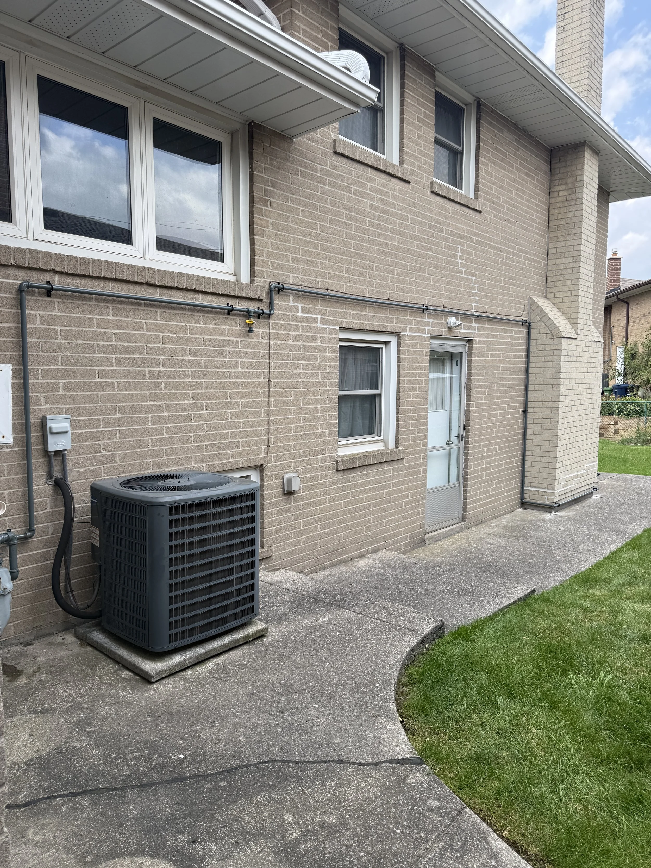 Backyard of a brick house with a concrete patio, green lawn, air conditioning unit outside, windows, door, and visible utility pipes.