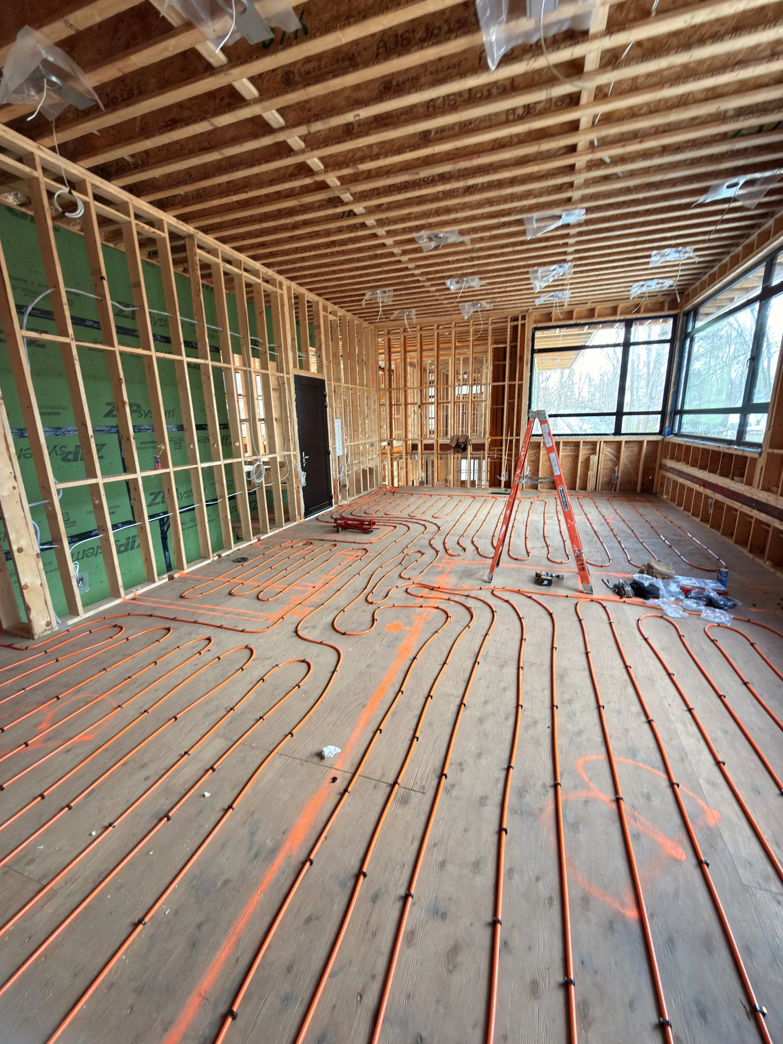 Under construction room with exposed wooden framing, orange plumbing pipes on the floor, and large windows on one side.