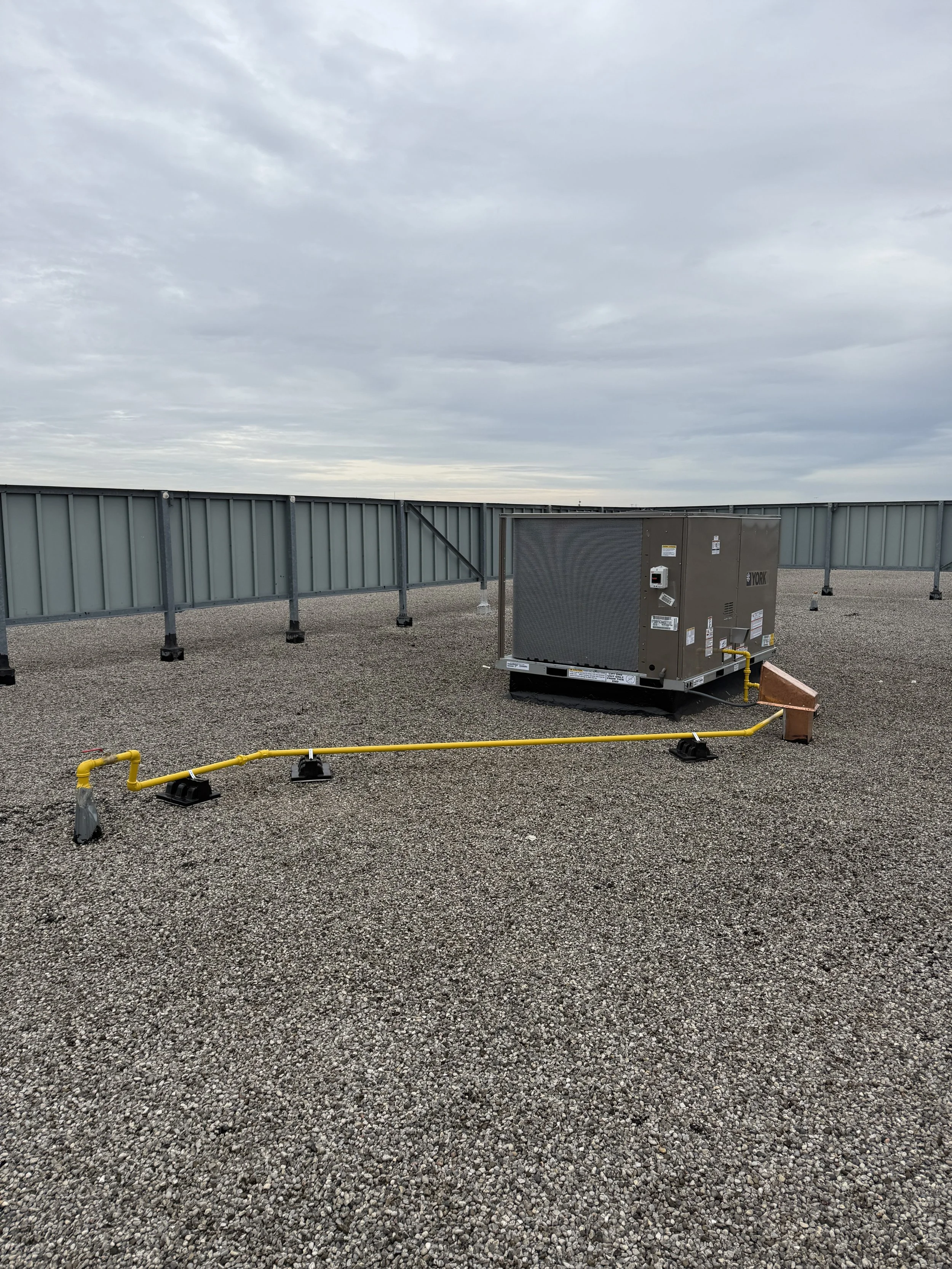 Rooftop with gravel surface, metal HVAC unit, and yellow gas line on the ground under a cloudy sky.