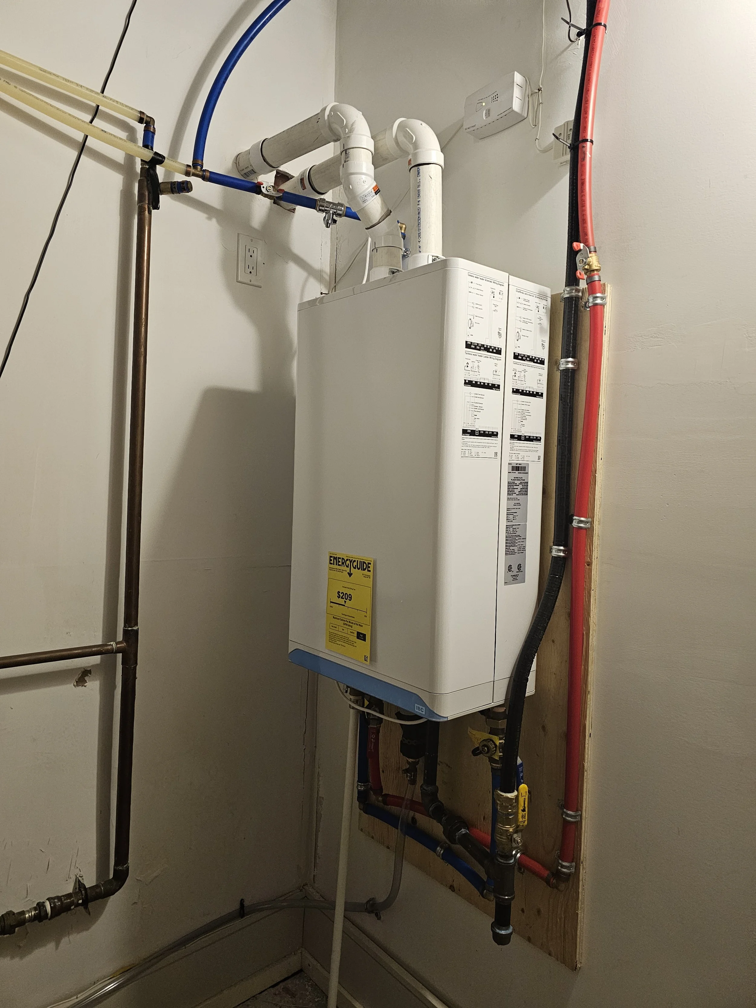A white tankless water heater mounted on a wooden board on a beige wall, with various pipes and fittings attached, including red, black, and copper piping, and blue and white plumbing lines. An energy guide label is visible on the front of the unit.