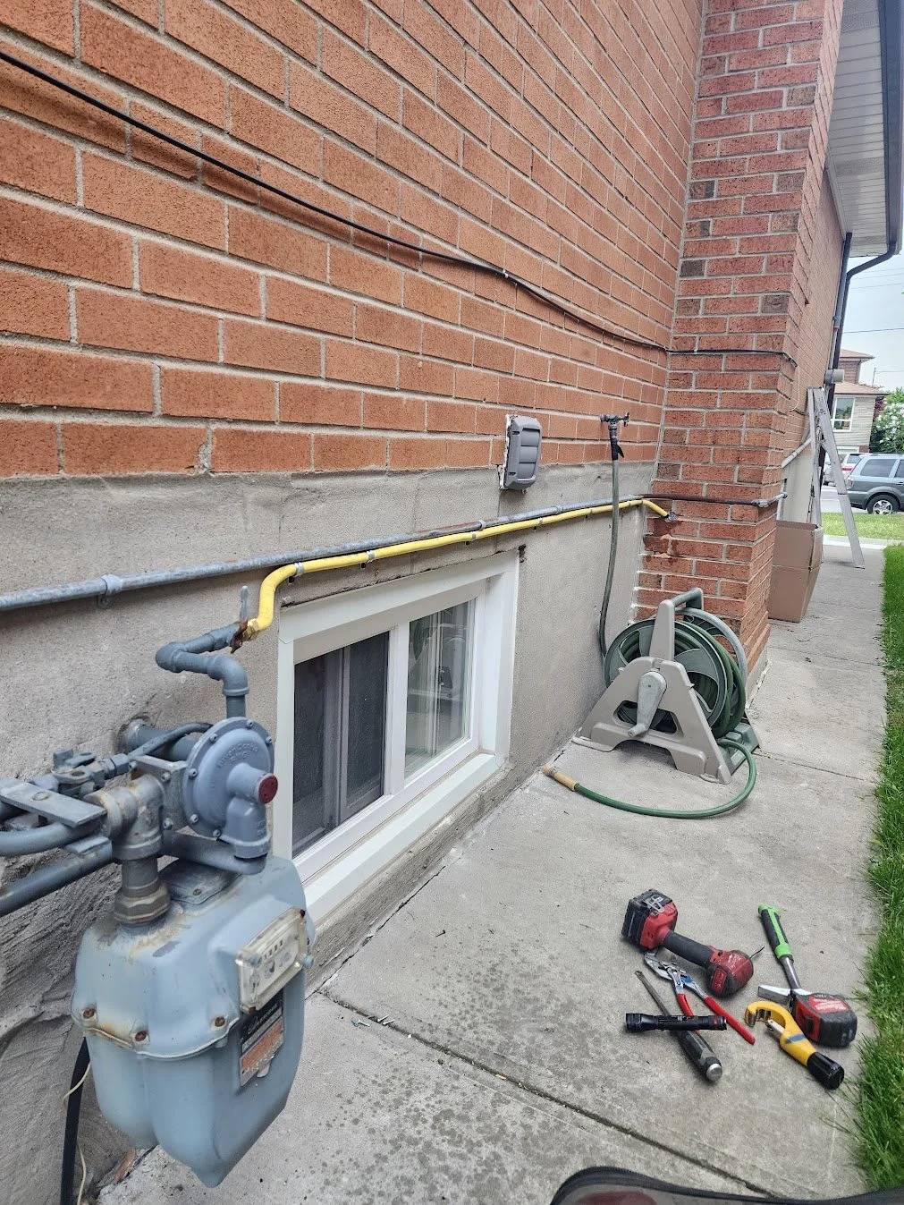 Side view of a brick house with construction tools and supplies for plumbing or siding work, including a water meter, a coiled garden hose, a figure with a ladder, and scattered tools on the concrete sidewalk.
