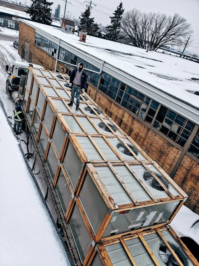 Worker standing on top of a large refrigeration unit mounted on a truck in a snowy urban environment, with another worker nearby.