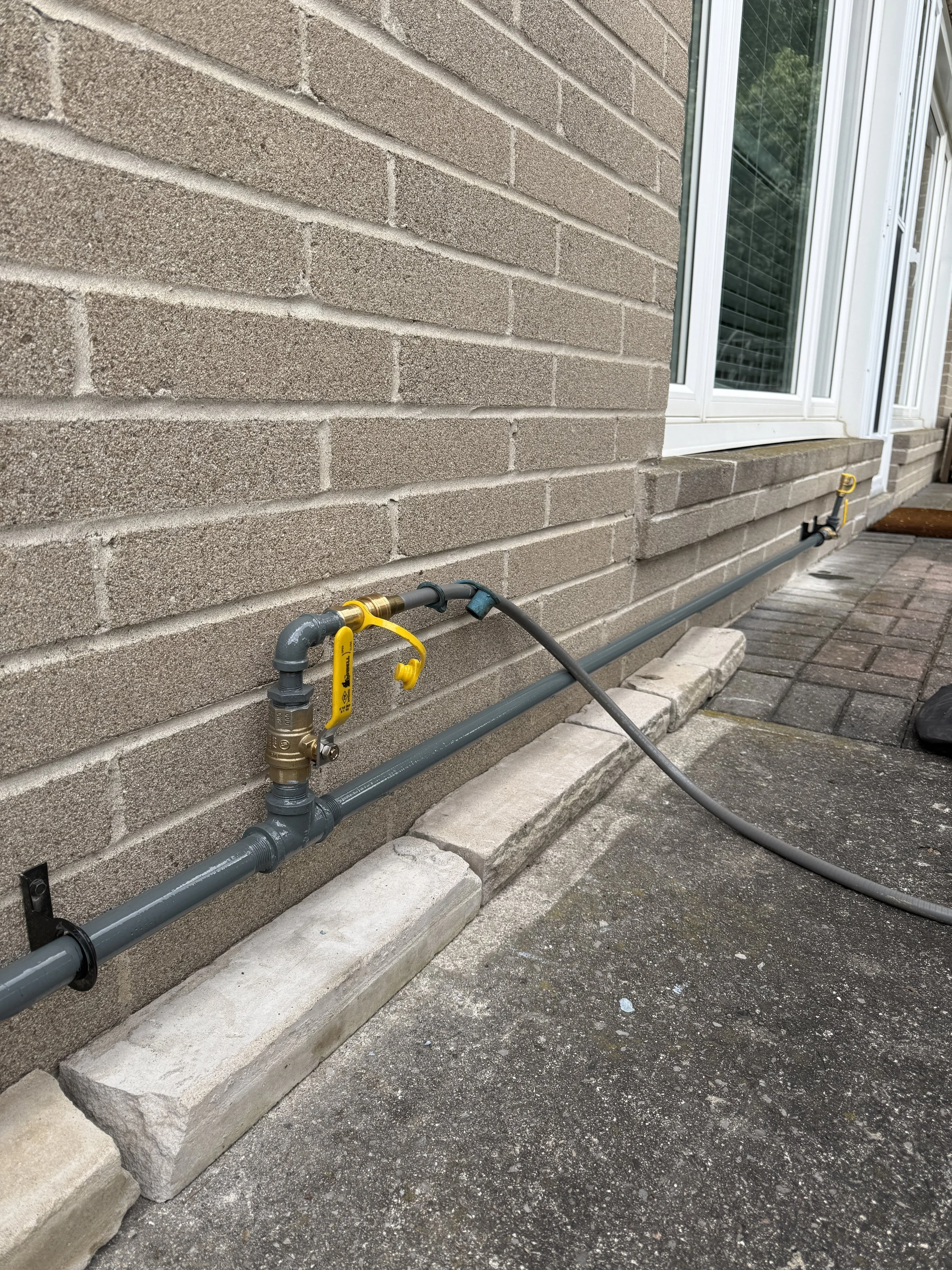 Exterior view of a house with a brick wall and a window, showing gray gas pipes running along the ground and attached to the wall, with yellow valve handles.