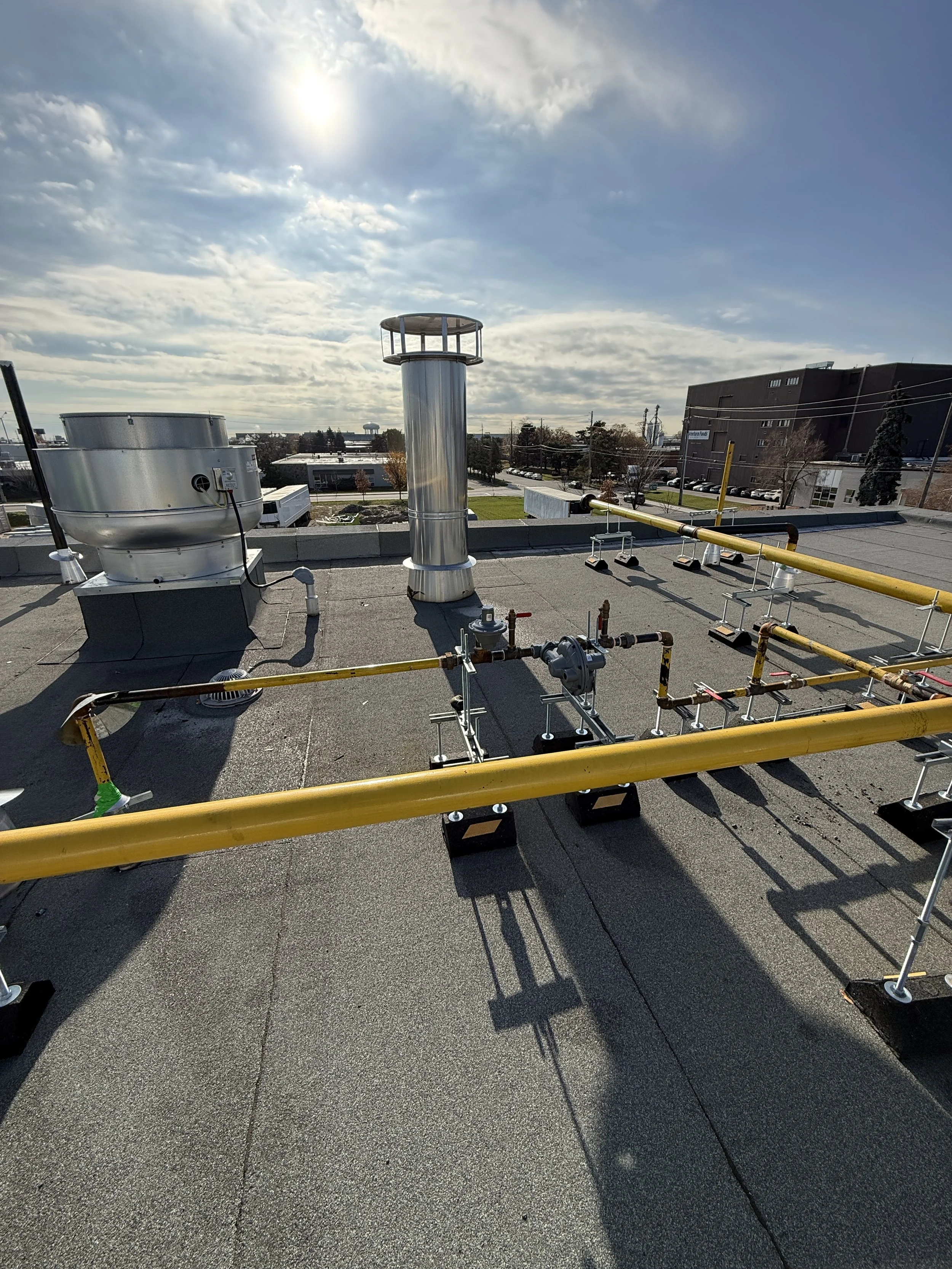 Rooftop with industrial pipes, vents, and a yellow gas line under a partly cloudy sky with sunlight.