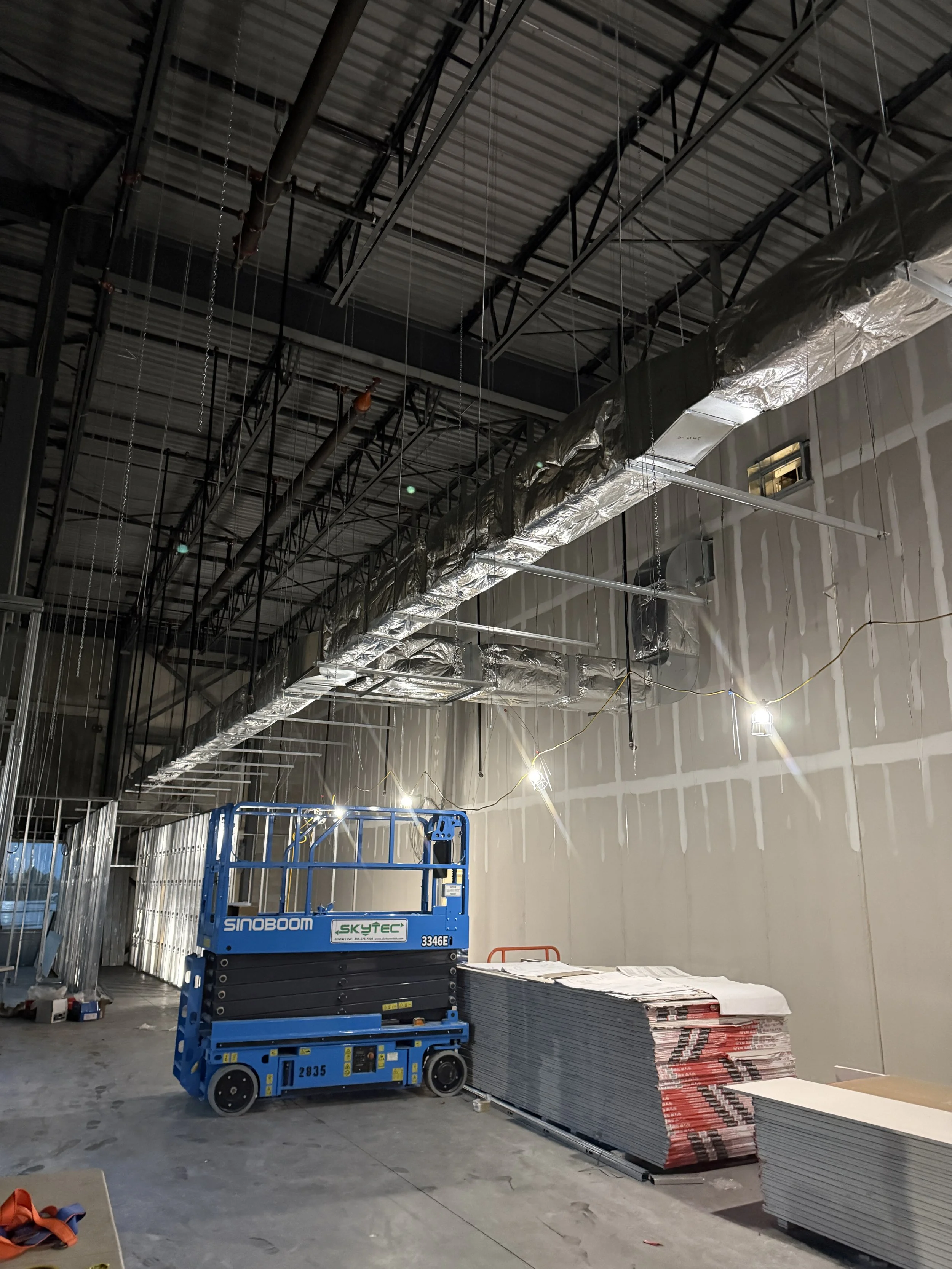 Construction site with scaffolding, ductwork, and construction materials including stacks of drywall and metal studs; a blue scissor lift is parked near the wall.