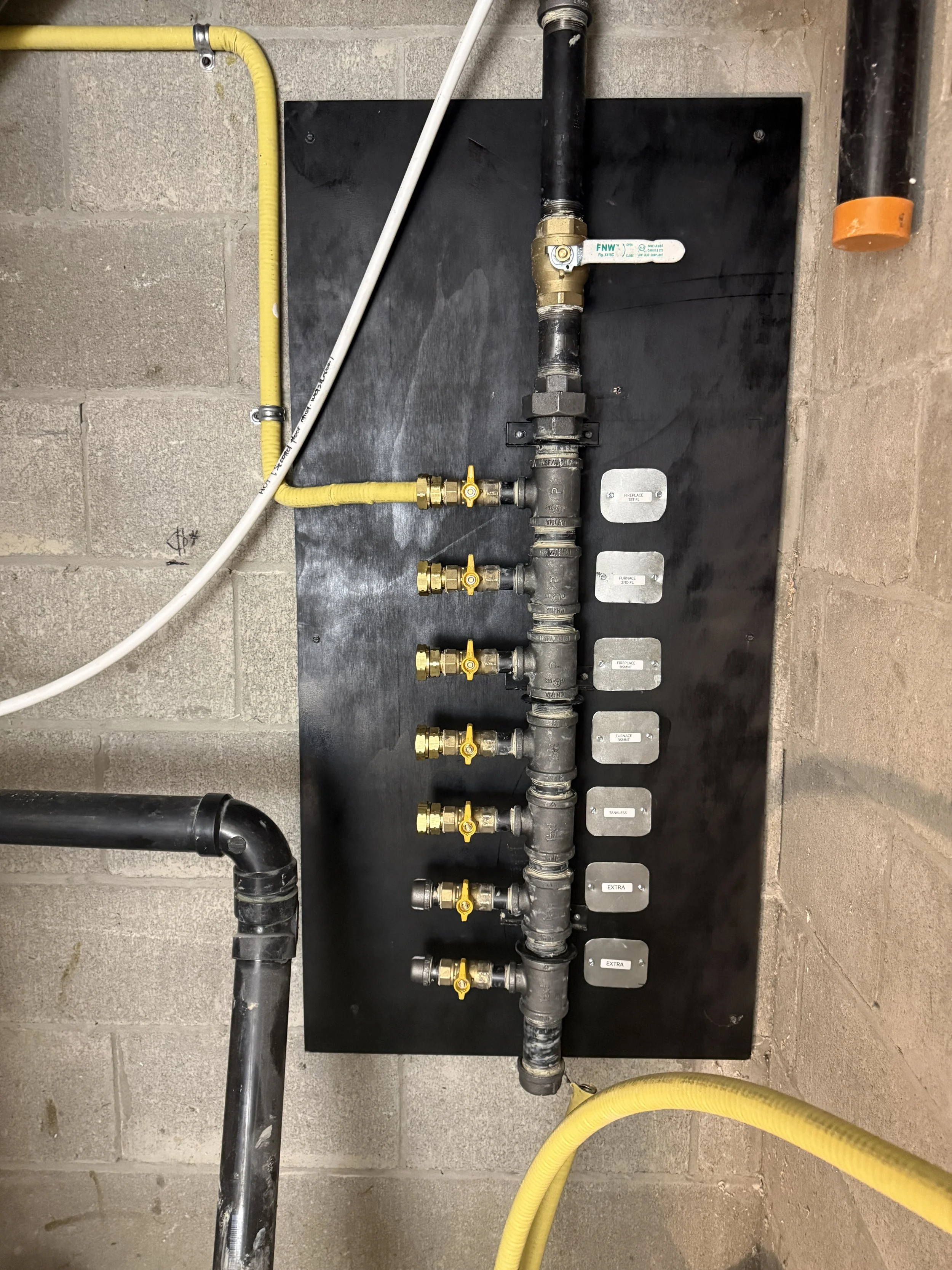 Industrial plumbing pipe system with multiple valves and labels mounted on a black panel against a concrete wall.