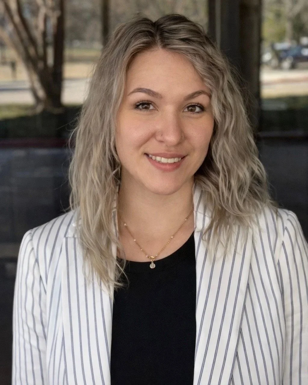 Paralegal Angela Roberts smiling, wearing a striped blazer and a black top, with a necklace and a nose piercing, indoors with a window and trees outside.