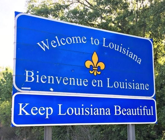 Blue road sign welcoming travelers to Louisiana with text in English and French, and a yellow fleur-de-lis symbol, reading 'Welcome to Louisiana,' 'Bienvenue en Louisiane,' and 'Keep Louisiana Beautiful.'
