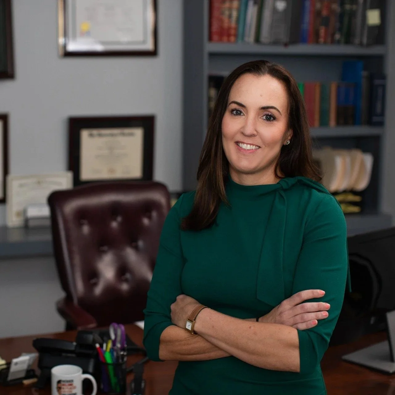 Attorney Lindsey Cheek in a green dress stands in an office, smiling with her arms crossed. Behind her are framed certificates and a bookshelf filled with books.