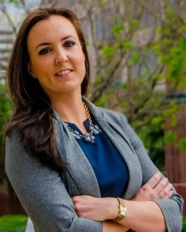Attorney Lindsey Cheek with a confident smile, wearing a gray blazer, blue top, and a statement necklace, standing outdoors with trees and greenery in the background.
