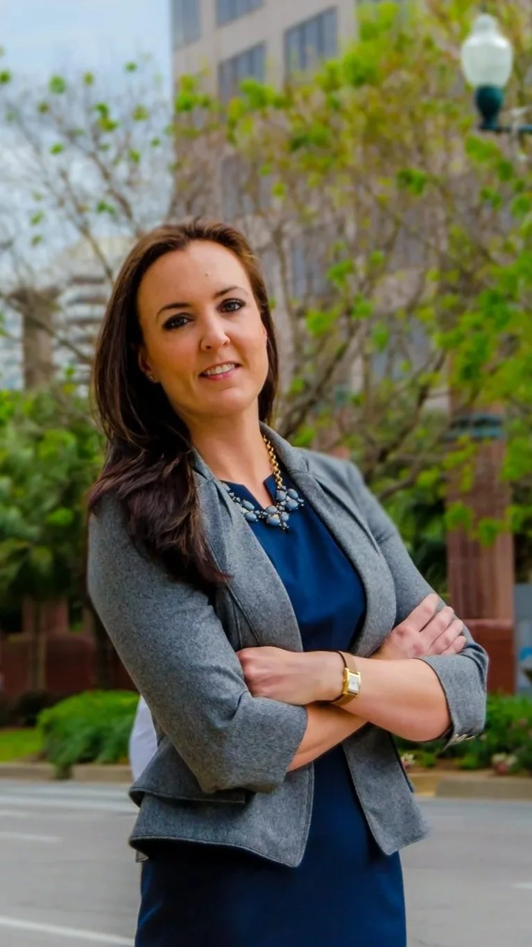 Paralegal Kelsey Cheek standing outdoors in front of trees and buildings, with her arms crossed, wearing a gray blazer, blue dress, and gold jewelry.