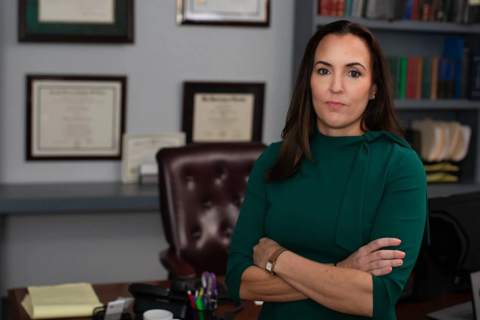 Attorney Lindsey Cheek in a green top standing with arms crossed in an office, with framed certificates on the wall and bookshelves in the background.