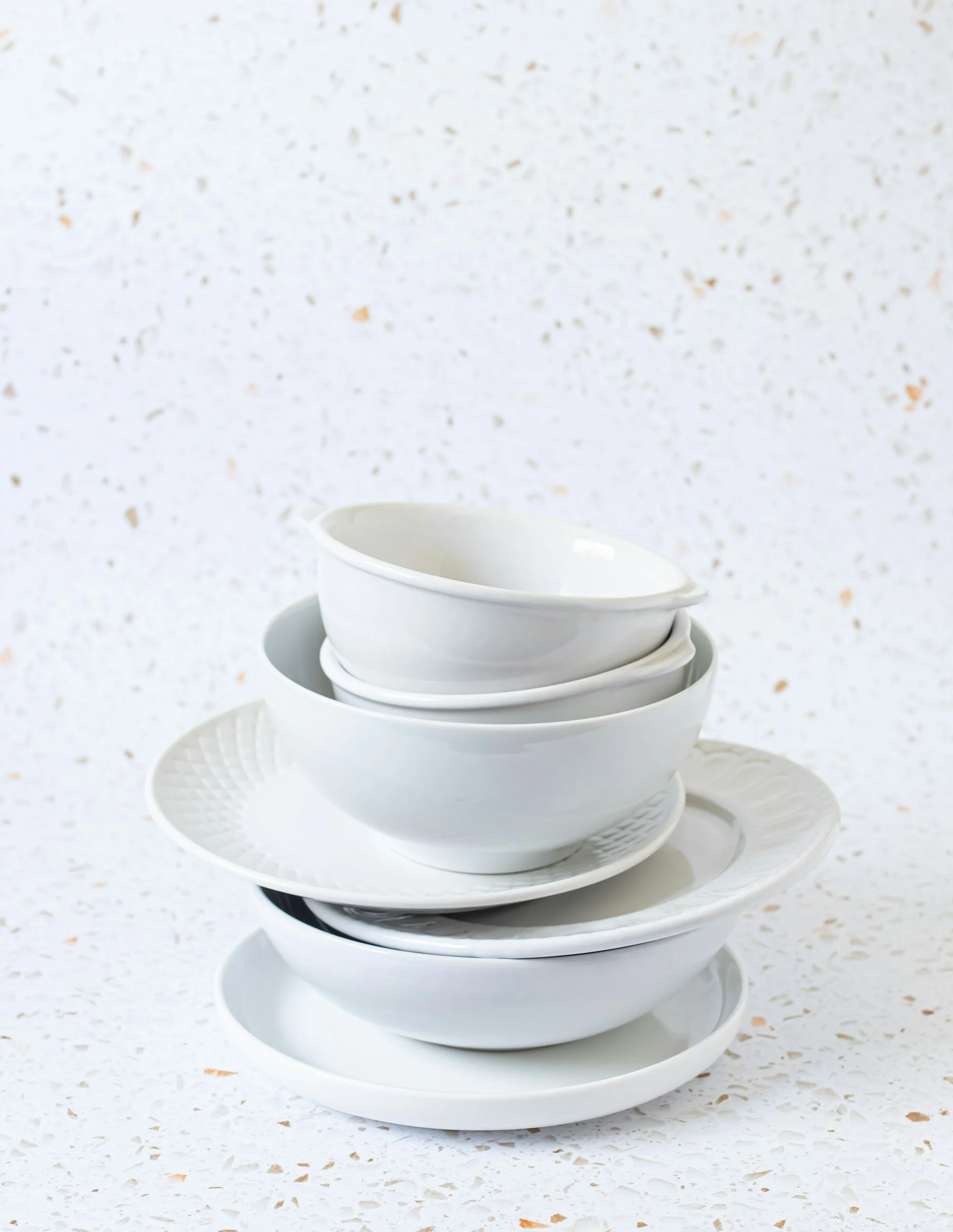 Stack of white ceramic bowls and plates on a speckled white background.