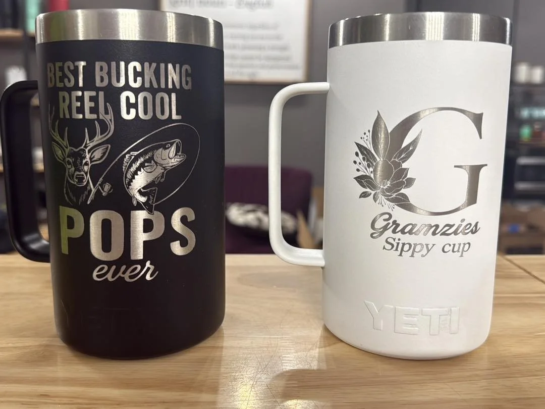 Two insulated coffee mugs on a wooden table; the left mug is black with green and white text and graphics, reading 'Best Bucking Reel Cool Pops Ever' with a deer and fish illustration; the right mug is white with silver text and design, showing a large letter 'G', floral accents, and the words 'Gramzies Sippy Cup', with a faint embossed 'YETI' logo at the bottom.